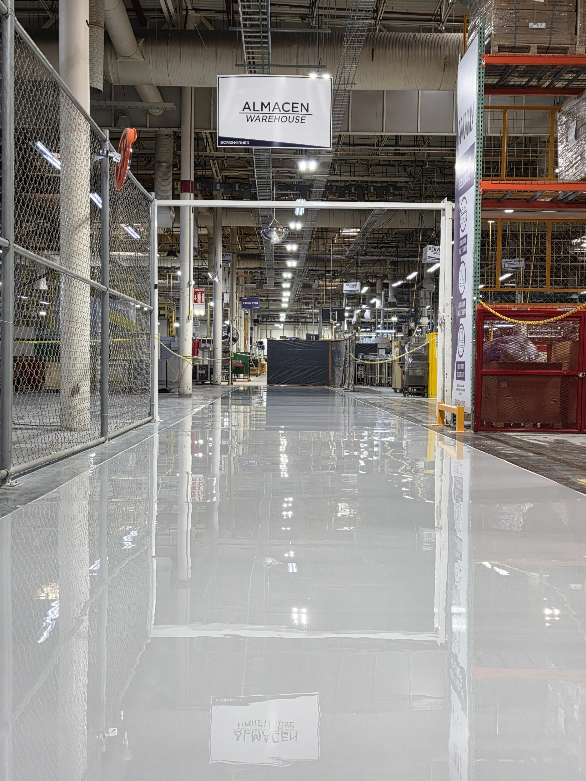Vista del interior de un almacén o fábrica con señal que dice "ALMACEN WAREHOUSE" y estructura metálica, estanterías y equipo industrial en el fondo.