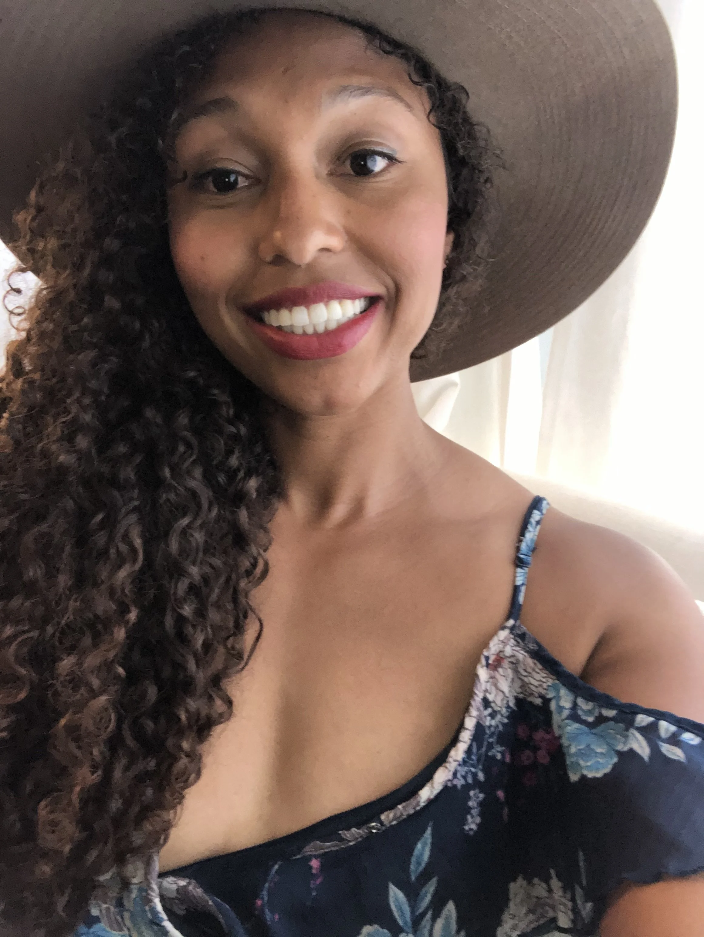 Close-up of a woman with curly hair, wearing a large wide-brimmed hat and a floral-patterned dress, smiling.
