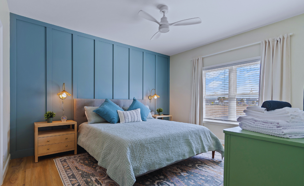 Primary king bedroom at Sol Mar with warm oak nightstands, a window with curtains and blue board and batten custom wall.  There are folded towels and linens on a green dresser.