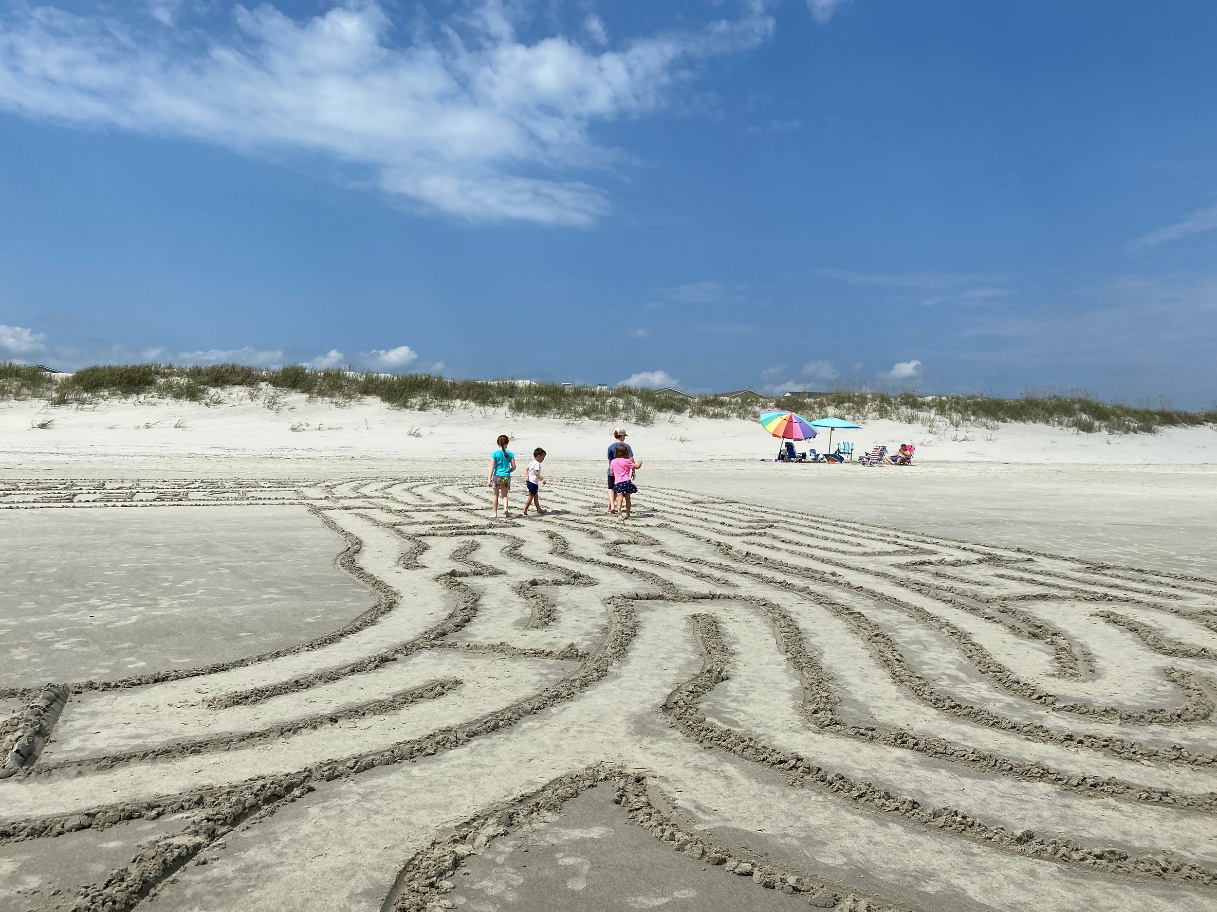 Kids walking along the beach maze in the sand, chairs and umbrellas in the distance.