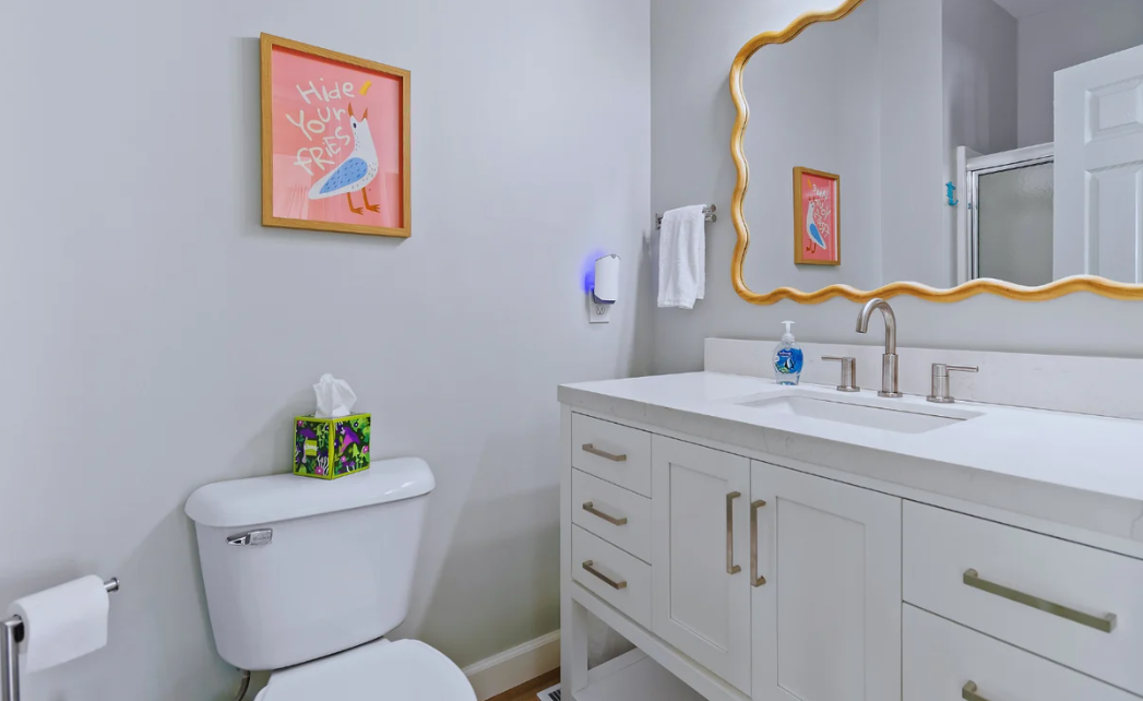 A bathroom at Sol Mar with light gray walls, a white toilet, a quartz topped white vanity with a mirror, a hand soap dispenser, a towel on a rack, and colorful framed artwork on the walls.
