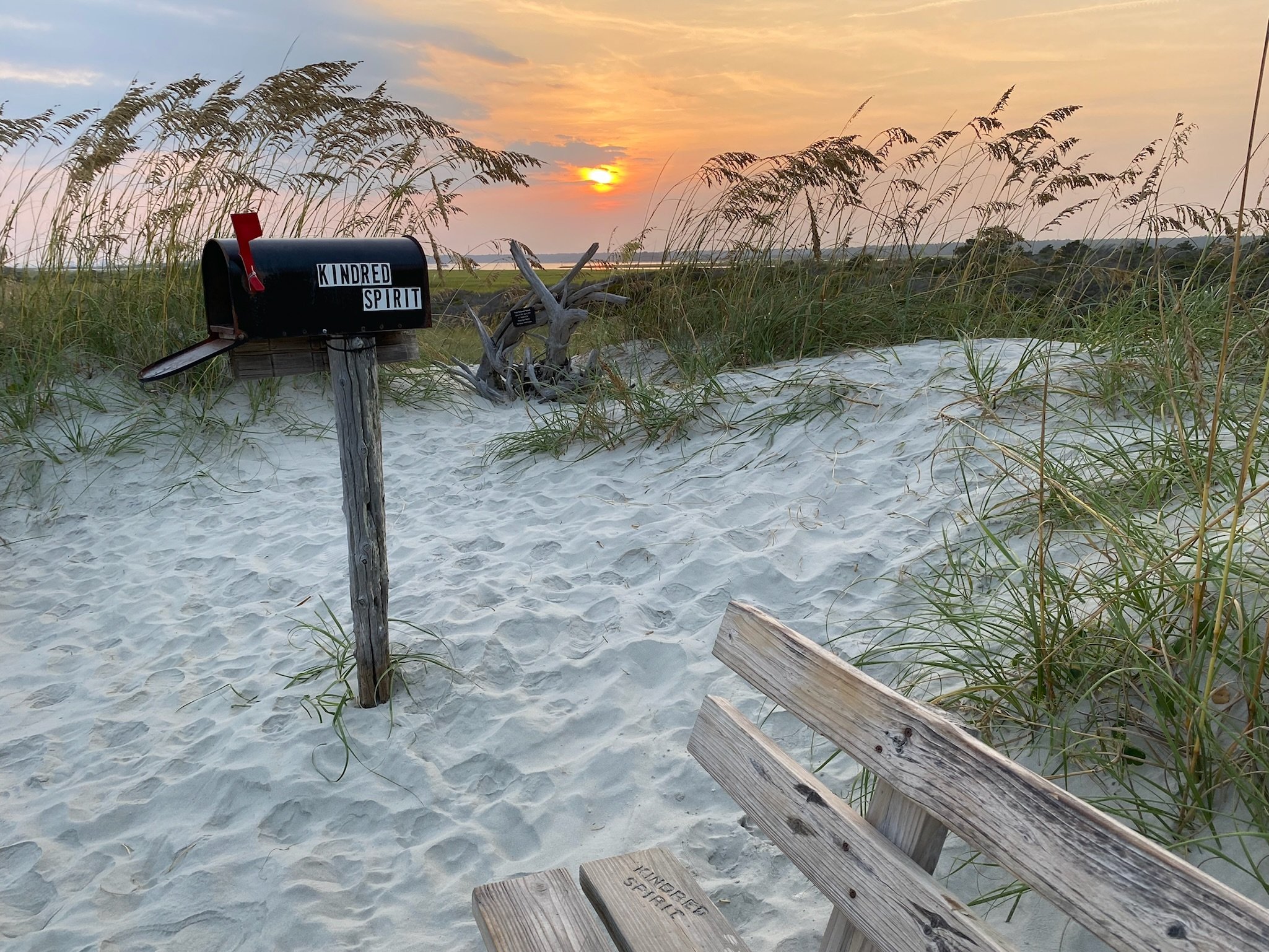 Kindred Spirit Mailbox on the beach at Sunset Beach, NC, with tall grass and a wooden bench at sunset.