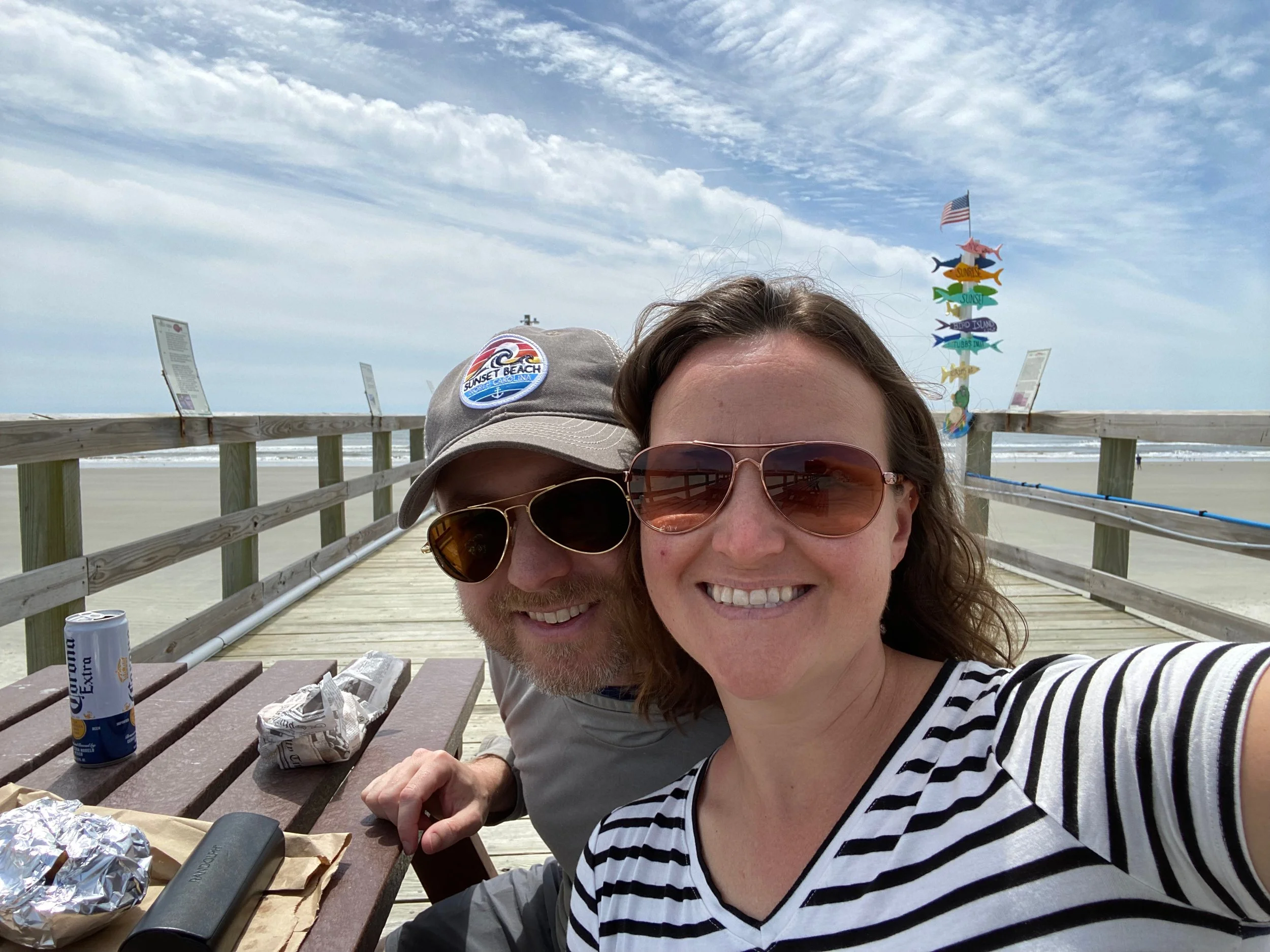 A smiling woman and a man taking a selfie at a beach pier, with rainbow-colored fish weather vanes and an American flag in the background. There are snacks, a can, and a speaker on a picnic table.