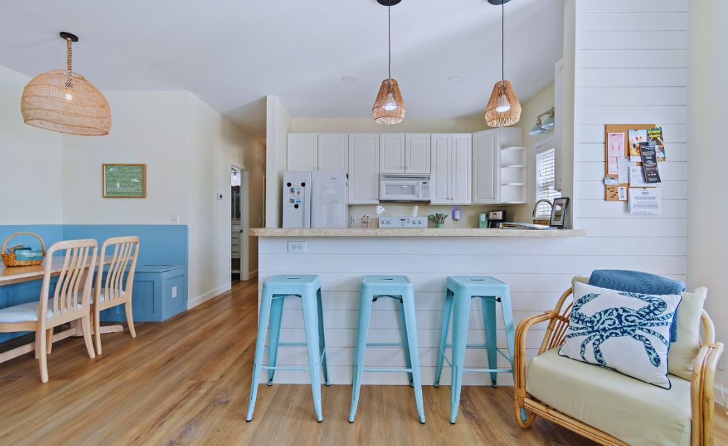 Kitchen at Sol Mar with white cabinets, light-colored countertops, three blue barstools, and a white wall with shiplap. Adjacent banquette dining area with a table, four chairs, and a wicker pendant light. A small sitting area with a wicker chair and