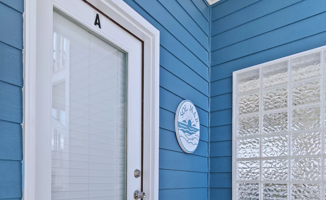 The exteriorentrnace of at Sol Mar with blue painted siding, a white door with a letter 'A' on top, a round sign that says 'SOL MAR' with a sun and waves illustration