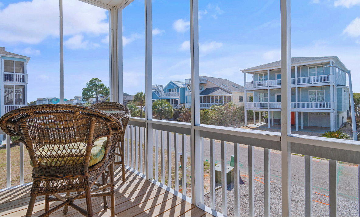 View from a screened porch at Sol Mar showing wicker chairs, neighboring houses with balconies, a sunny blue sky, and palm trees.