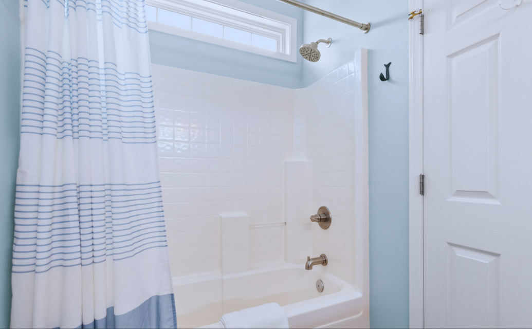 Primary bathroom shower at Sol Mar with white tiles, a small window, a rain shower head, a metal handle, a bathtub with a towel, light blue walls, and a white door.