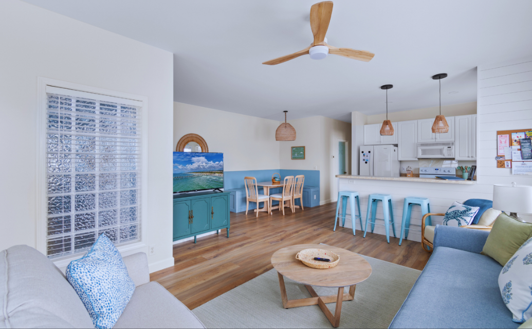 Open-concept living room at Sol Mar with white walls, light-colored sofa with patterned pillows, a wooden coffee table, and a green TV cabinet with a flat-screen TV. A banquette dining area with a wooden table and chairs, a white kitchen with hanging