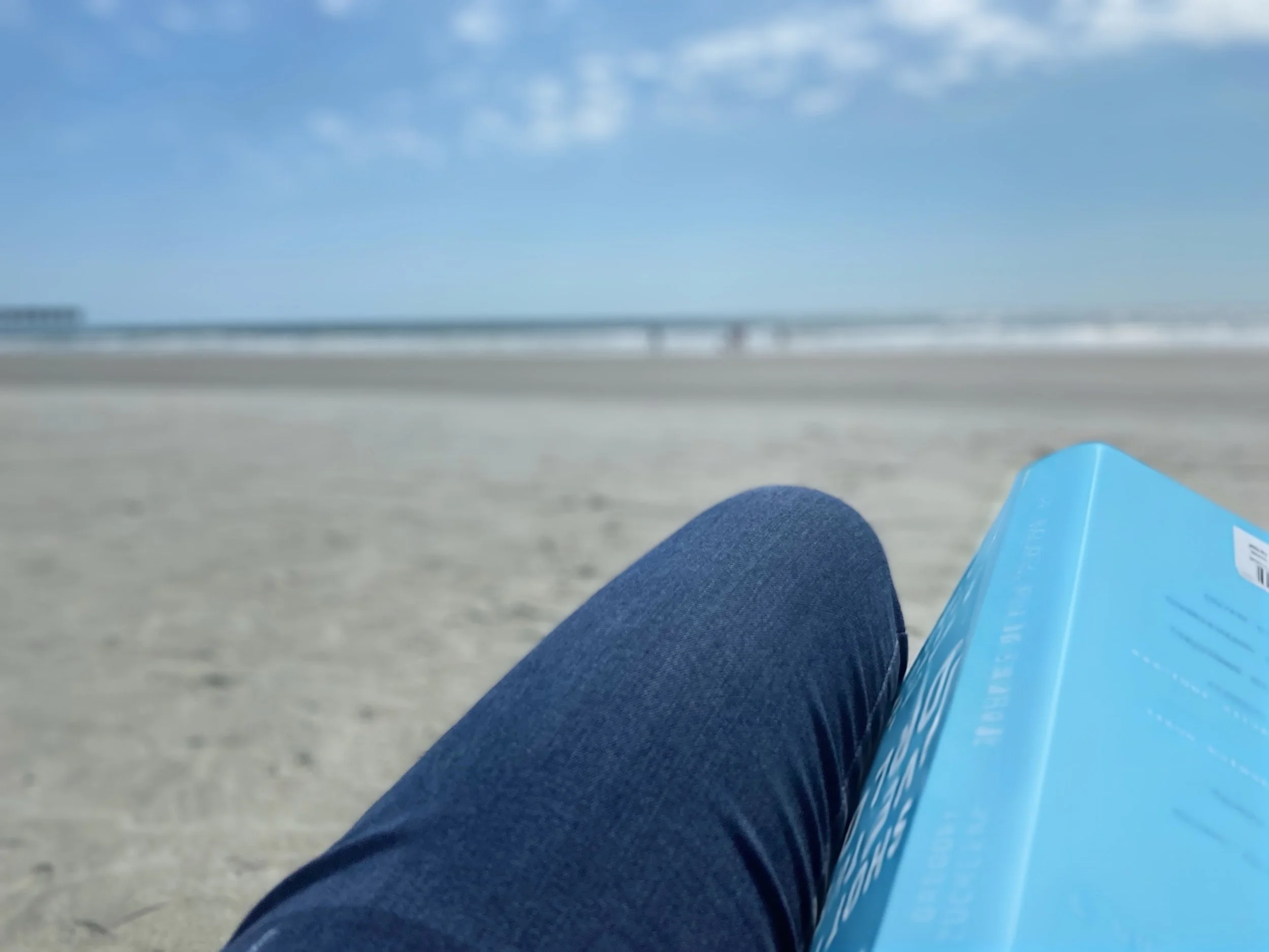 Person relaxing on the sand at Sunset Beach with a beach book with nearby ocean in the background.