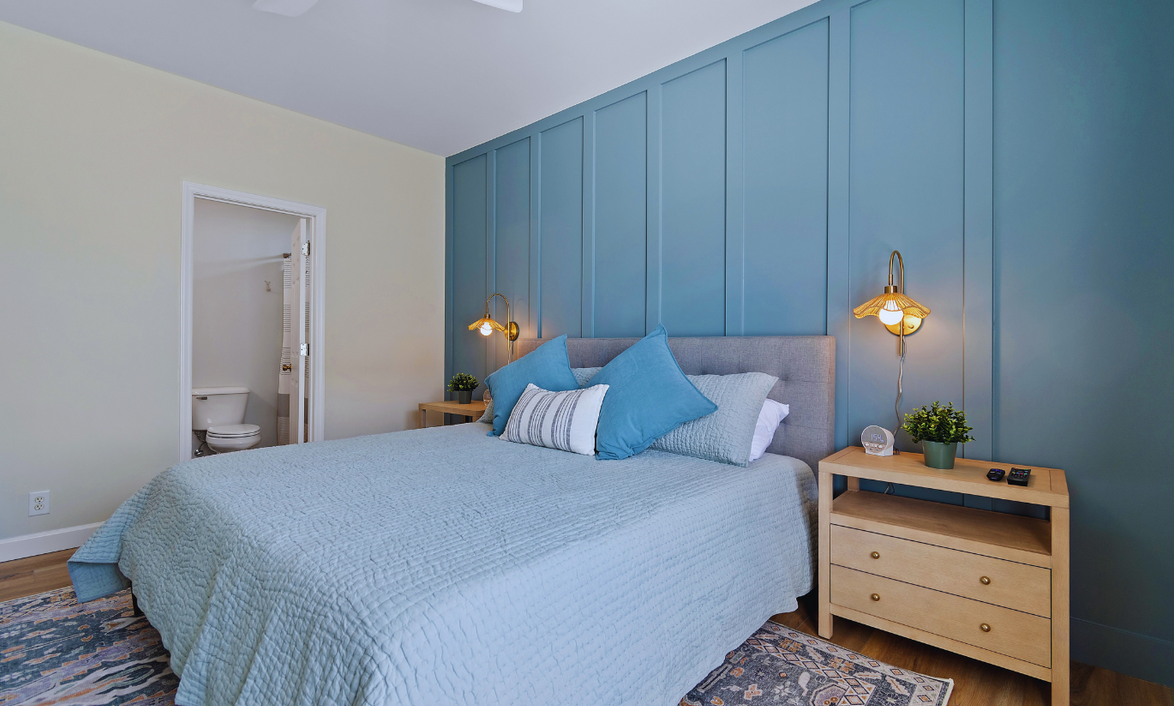 A primary king bedroom at Sol Marwith a bed, blue pillows, a blue accent wall, nightstands with lamps and plants, and a view of a small bathroom with a toilet.