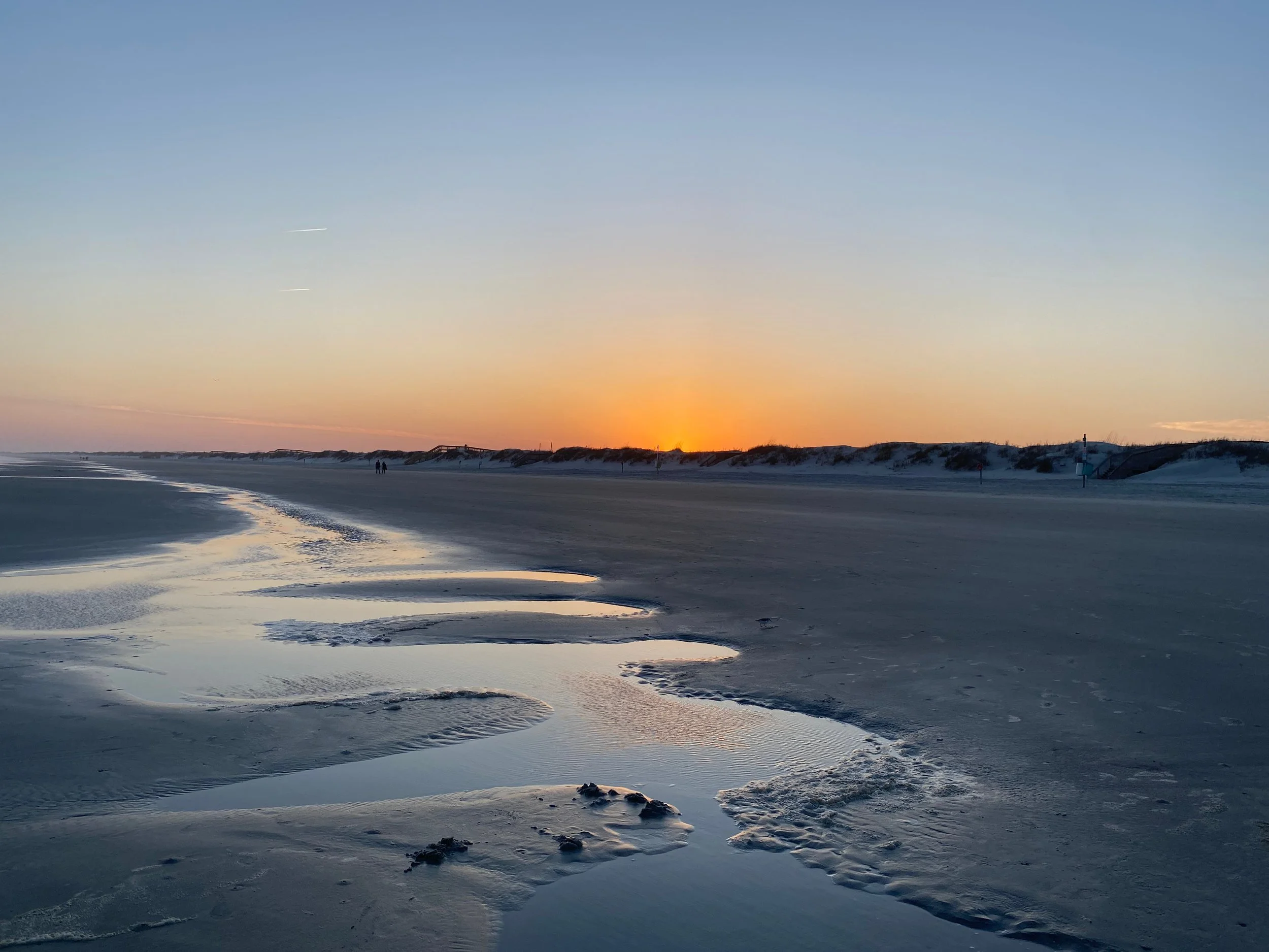 Beach at sunset with tide pools, sand dunes in the background, and a couple walking near the shoreline.