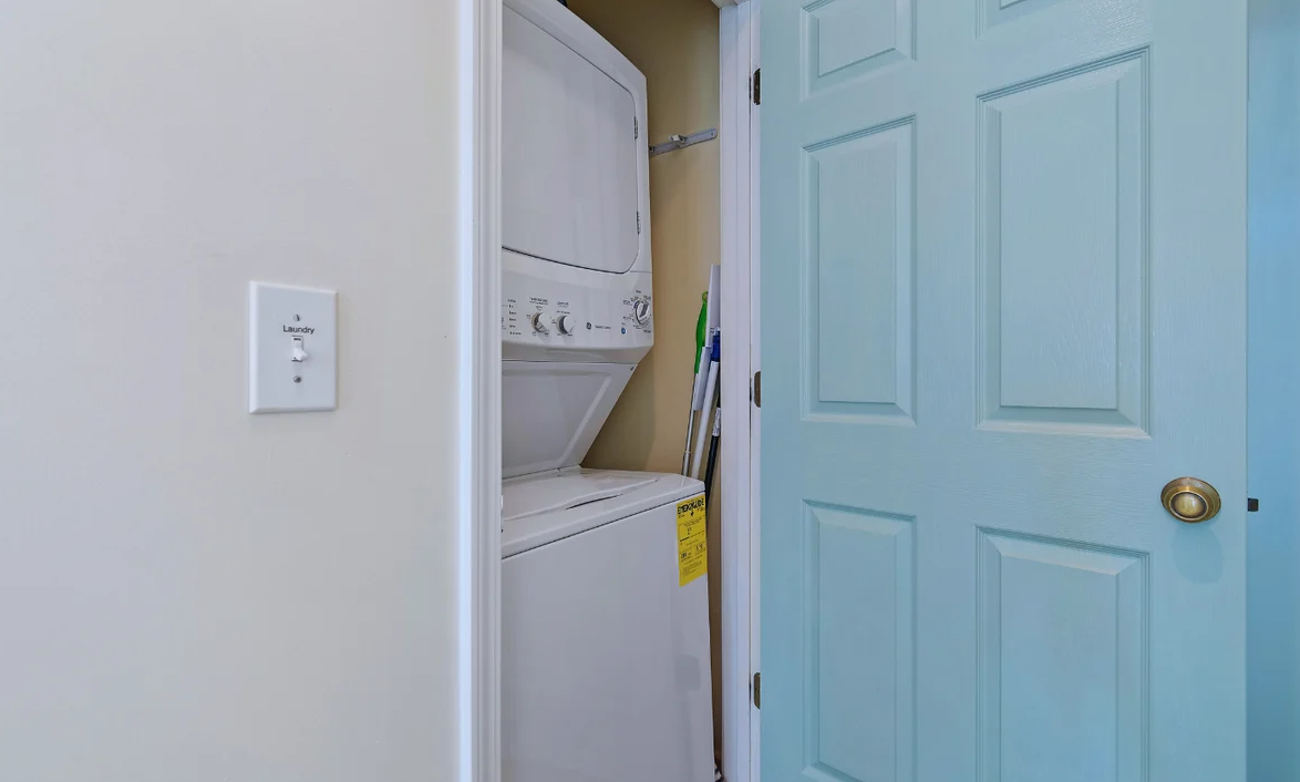 A small laundry closet at Sol Mar with a stacked washer and dryer, a broom and a mop with a blue door.