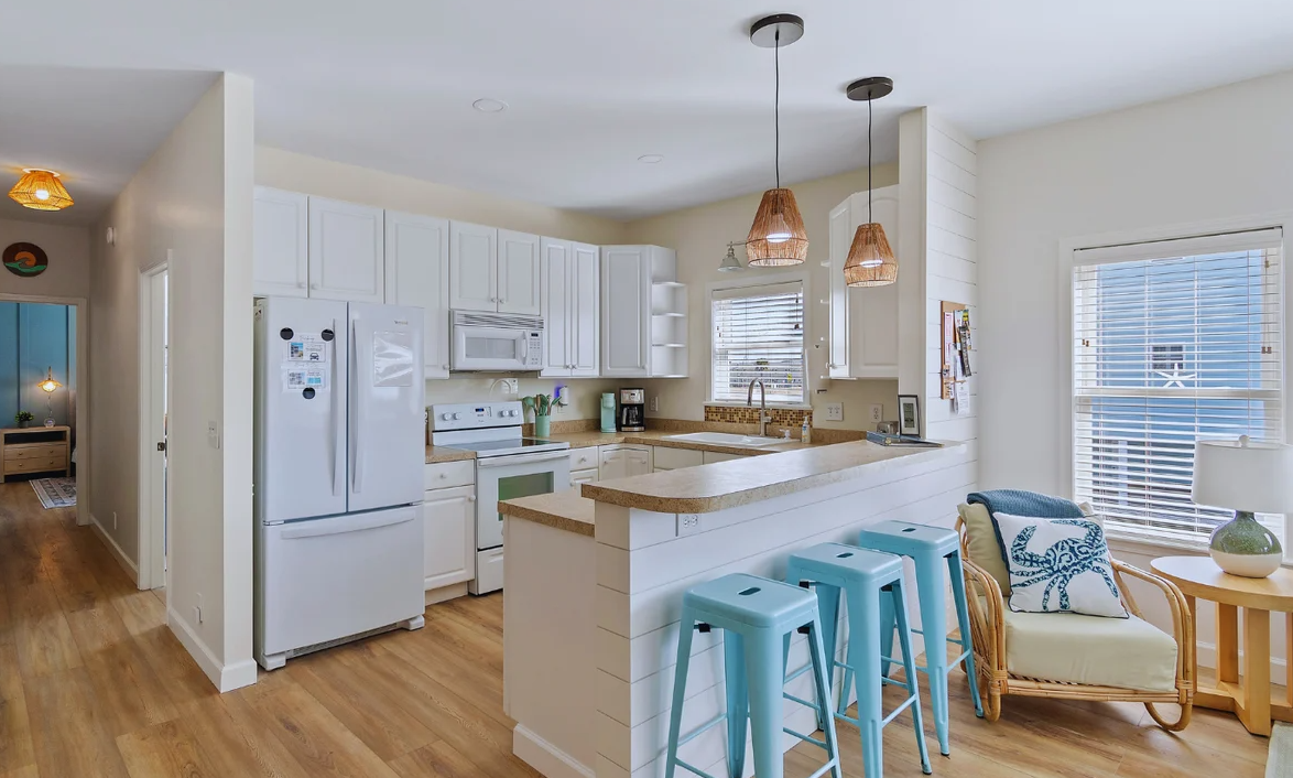Bright kitchen at Sol Mar with white cabinets, beige countertops, and wooden flooring, featuring hanging pendant lights and a small sitting area with a wicker chair, side table, and large windows.