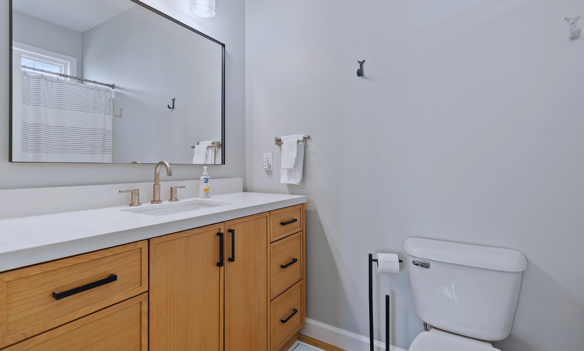 Primary bathroom at Sol Mar with a large mirror, wooden vanity with black handles, white quartz countertop, brushed gold faucet, soap dispenser, and a white toilet with black accessories. Light gray walls, window with striped curtain, and hooks on th