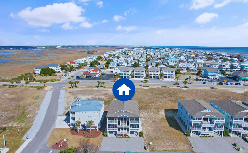 Aerial view of a coastal neighborhood with Sol Mar condo building and apartment complexes near the beach, featuring a large blue icon with a white house symbol in the center.