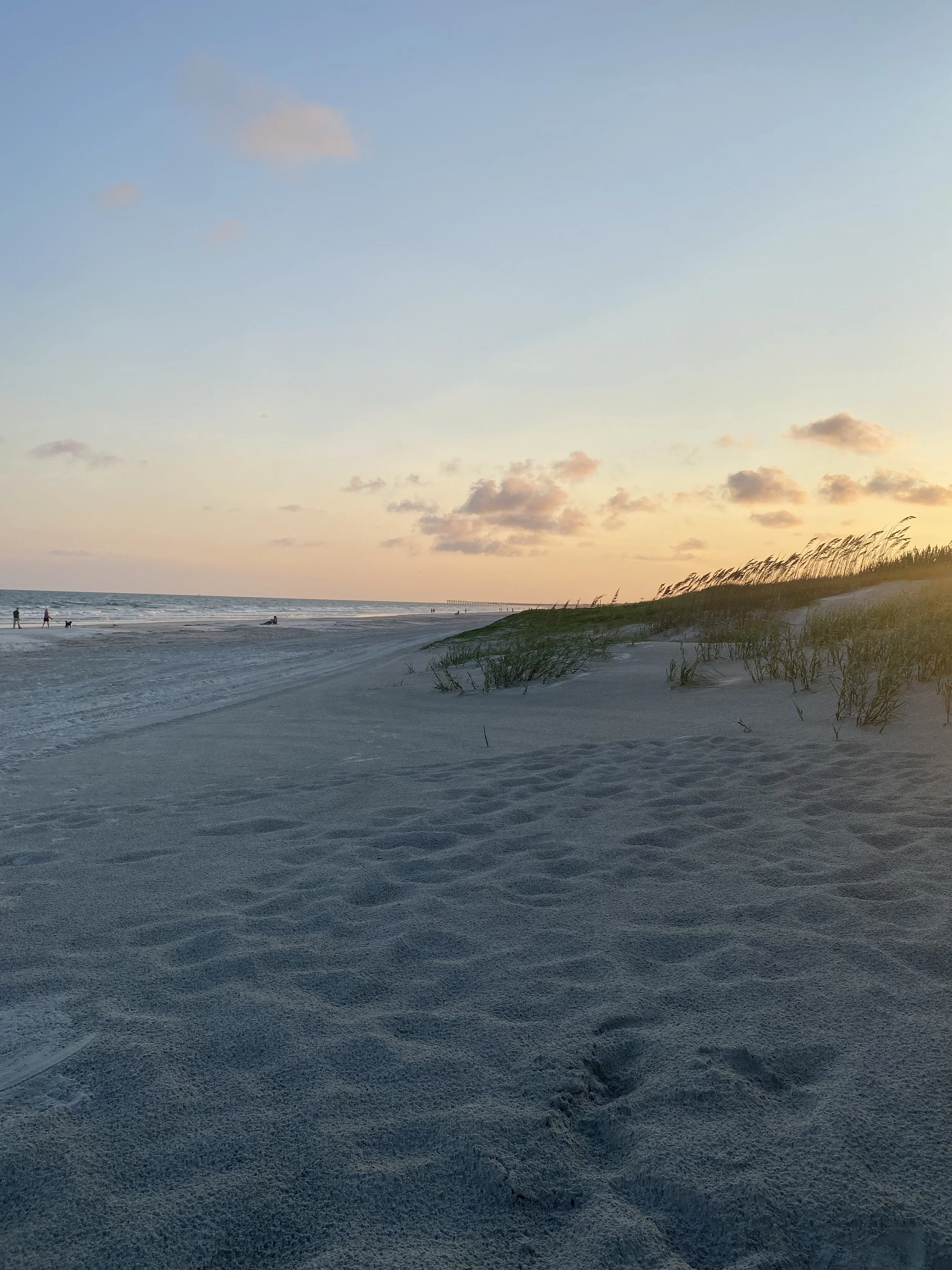 Golden hour view of the dunes and shoreline at Sunset Beach, North Carolina, with sea oats blowing in the foreground and people walking the beach in the distance.