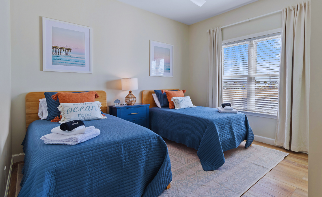 Second bedroom at Sol Mar with two twin beds in navy bedding, orange accent pillows, and beach-themed wall art.