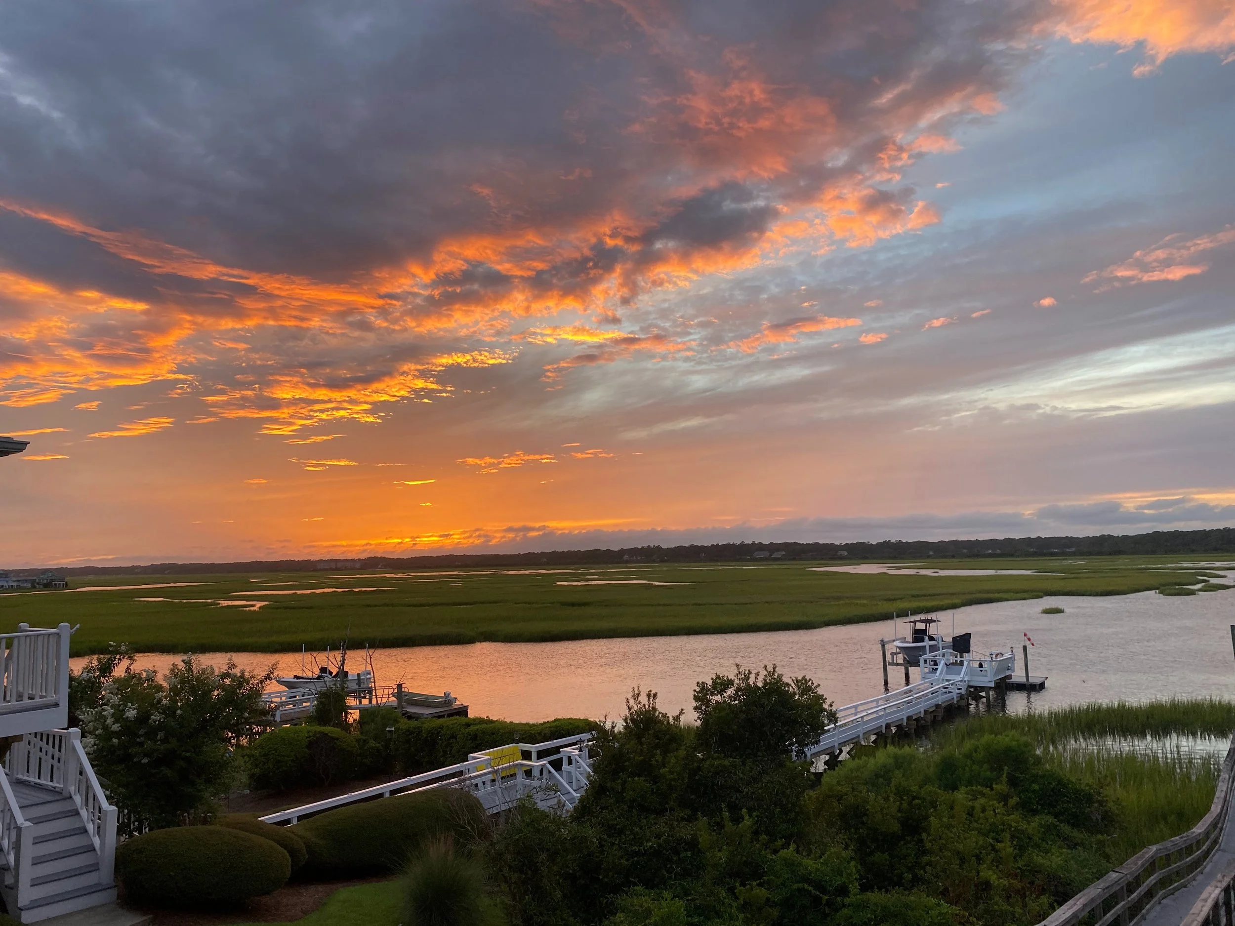 A scenic view of a sunset sky with orange, pink, and purple clouds over a river or wetland area. There are boats docked at a pier or dock in the foreground, and a white railing along the water's edge. Trees and shrubs are visible near the dock, with a residential house partially visible on the left.