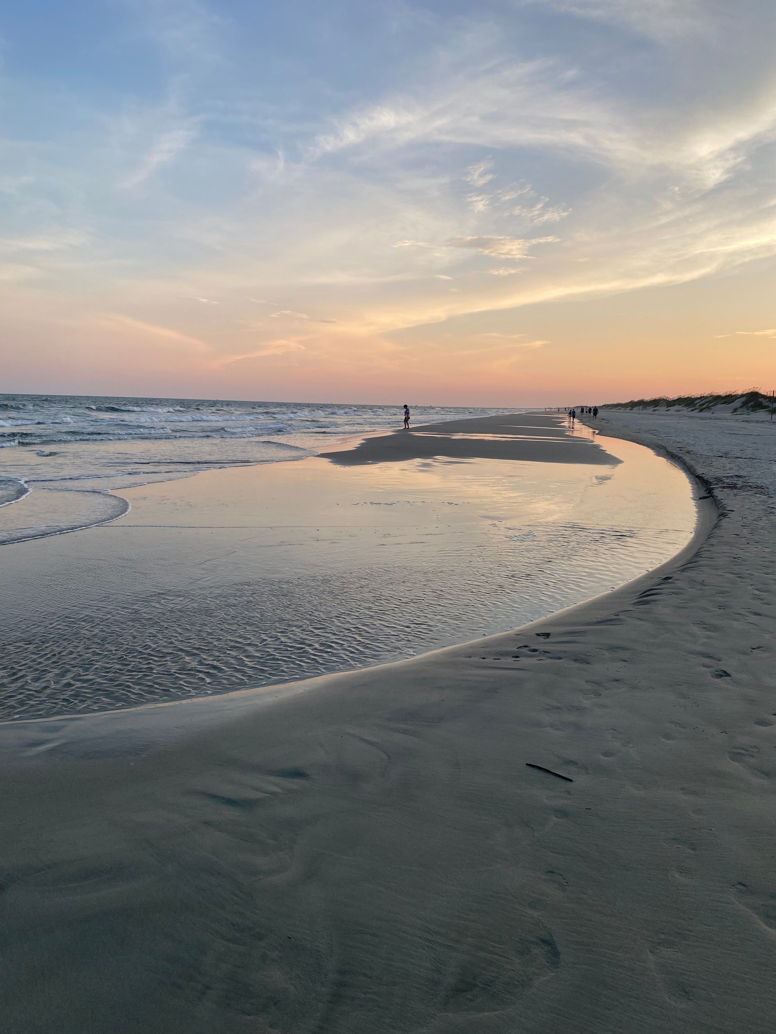Low tide reflection pool on the wet sand at Sunset Beach, North Carolina, with a pink and gold sunset sky and people walking along the shoreline.