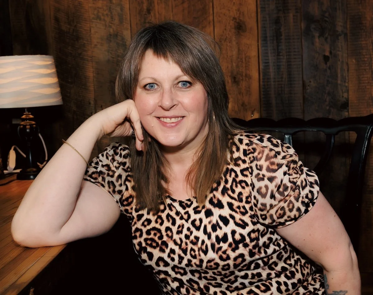 Laura, a white woman with blue eyes and a cute asymmetrical shag and bangs, wears a leopoard print crop top and sits on a vintage chair. She is smiling and resting her face on her hand.  Photo by Sarah Elizabeth Larson