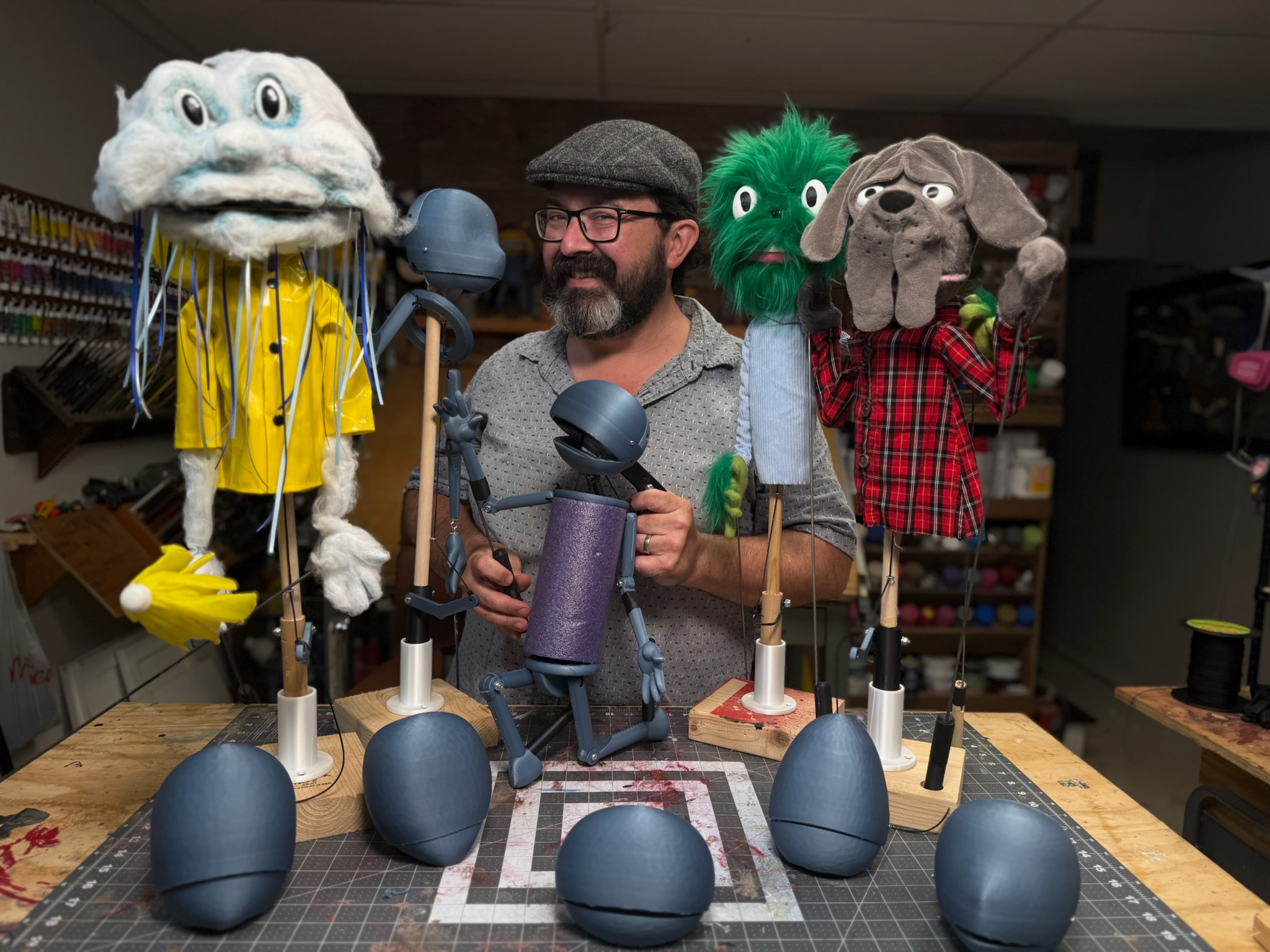 A man with a beard and glasses wearing a gray cap surrounded by puppets with expressive faces in a workshop. The puppets include a large white face with mustache and blue eyes, a bright green face with a beard, and a brown face with floppy ears. They are all held up on sticks and appear to be part of a puppet show or puppet-making project.
