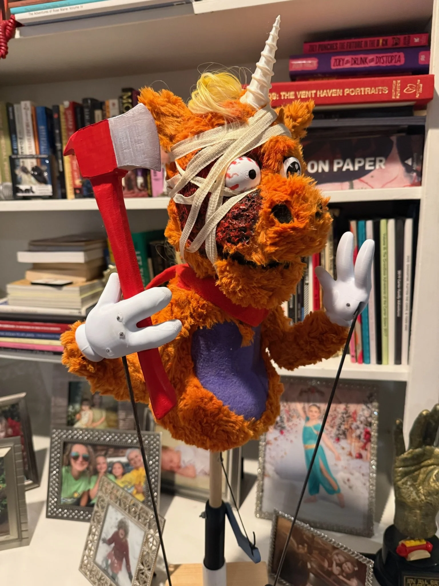 A plush orange monster puppet with a horn, large googly eyes, and a fuzzy mane holds a red axe in one hand. It is in front of a bookshelf with various books and framed photos, with other small objects and decorations in the scene.