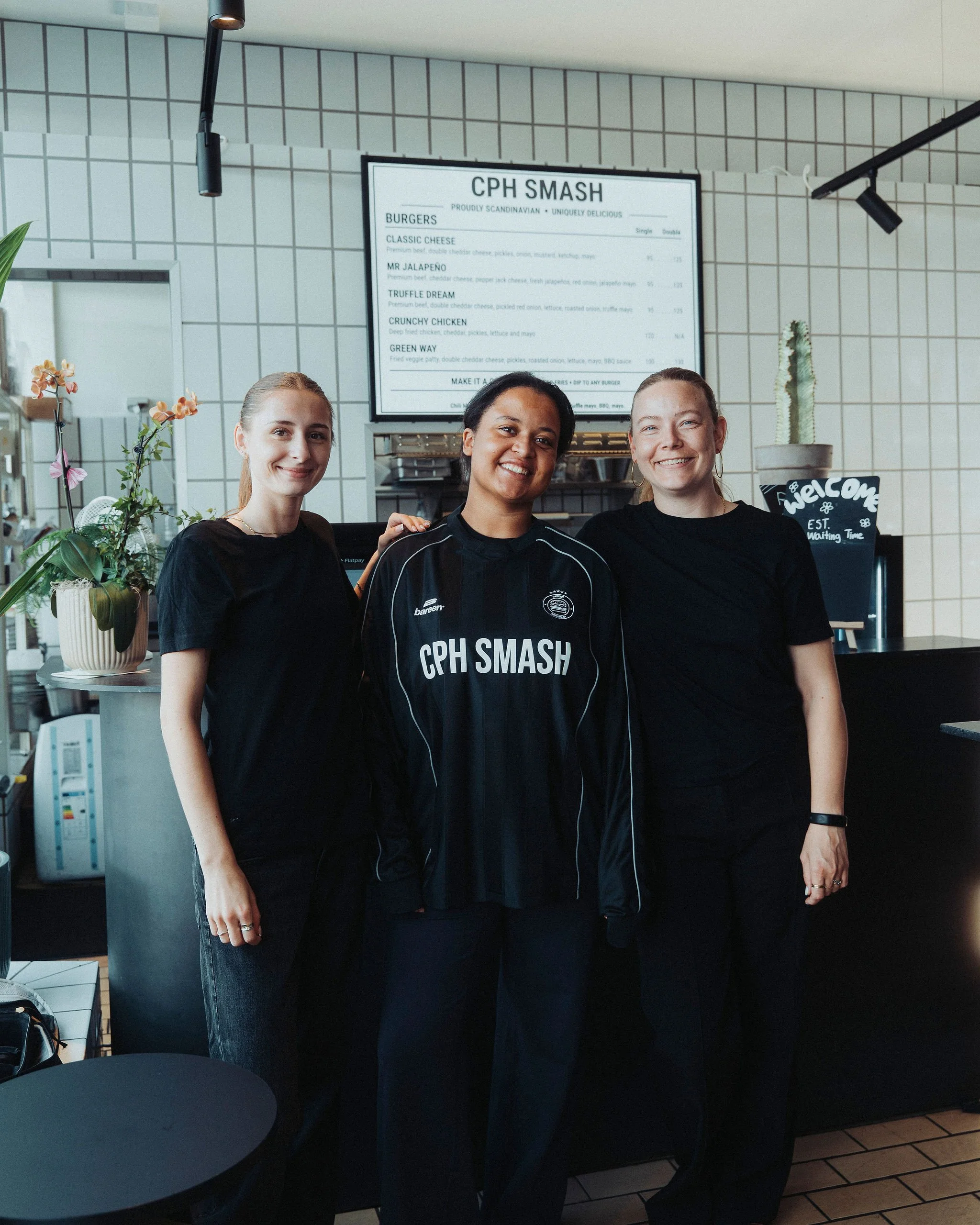 Three women standing inside a cafe, smiling at the camera, with a menu board behind them and potted plants on the counter.
