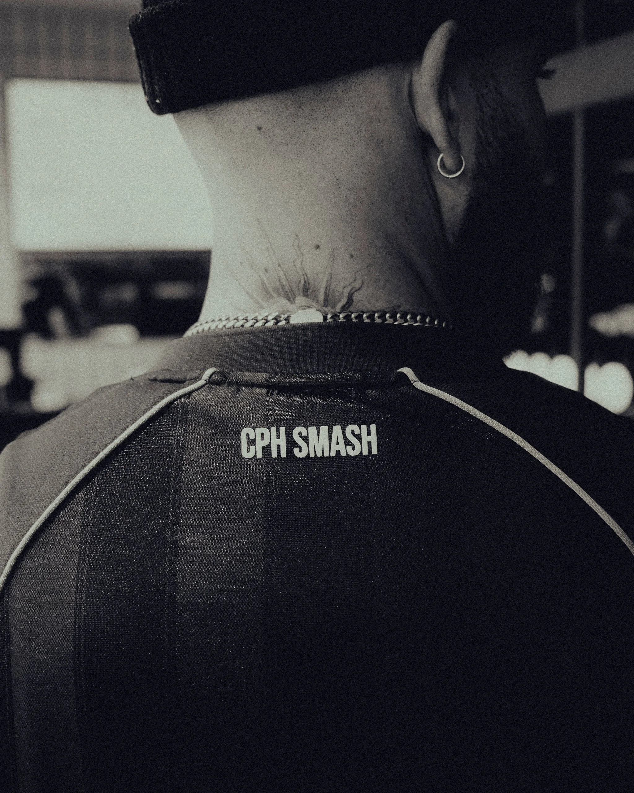 The back of a person's head with short dark hair, earrings, a tattoo on the neck, and wearing a black shirt with white text reading 'CPH SMASH'.