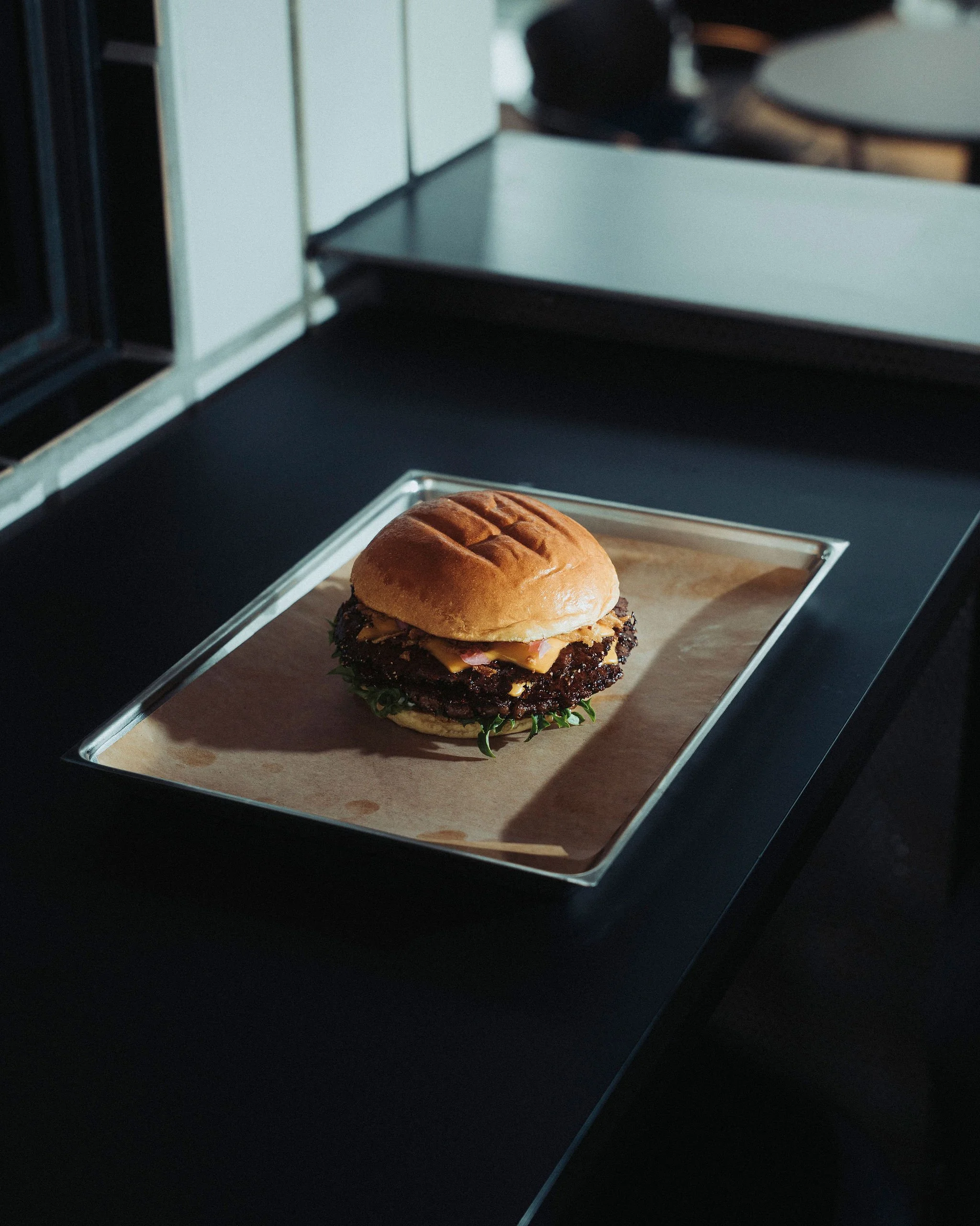 A burger with a bun, lettuce, tomato, cheese, and a beef patty on a wooden tray, with a black kitchen counter and cabinets in the background.