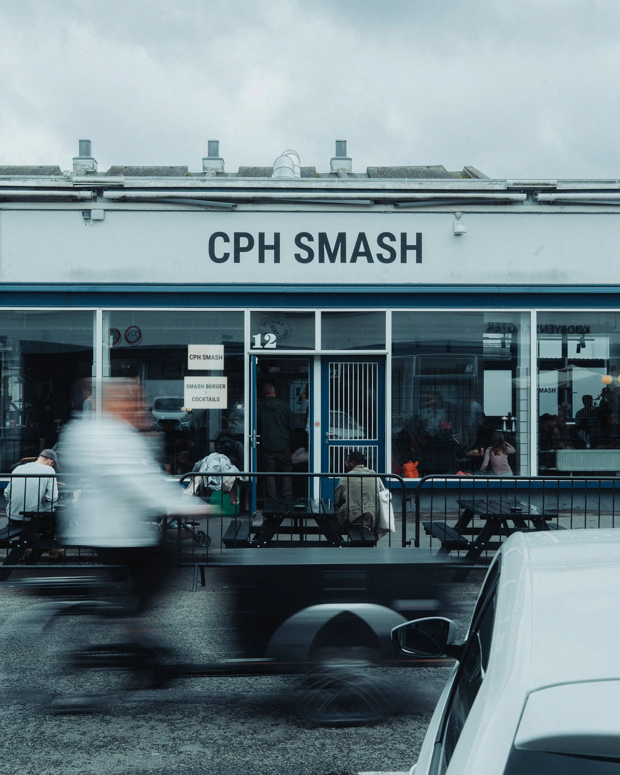 View of a fast food restaurant named CPH Smash with customers sitting outside, metal picnic tables, and a parking lot with cars. The restaurant has large glass windows and a sign indicating they serve smash burgers, cocktails, and more.