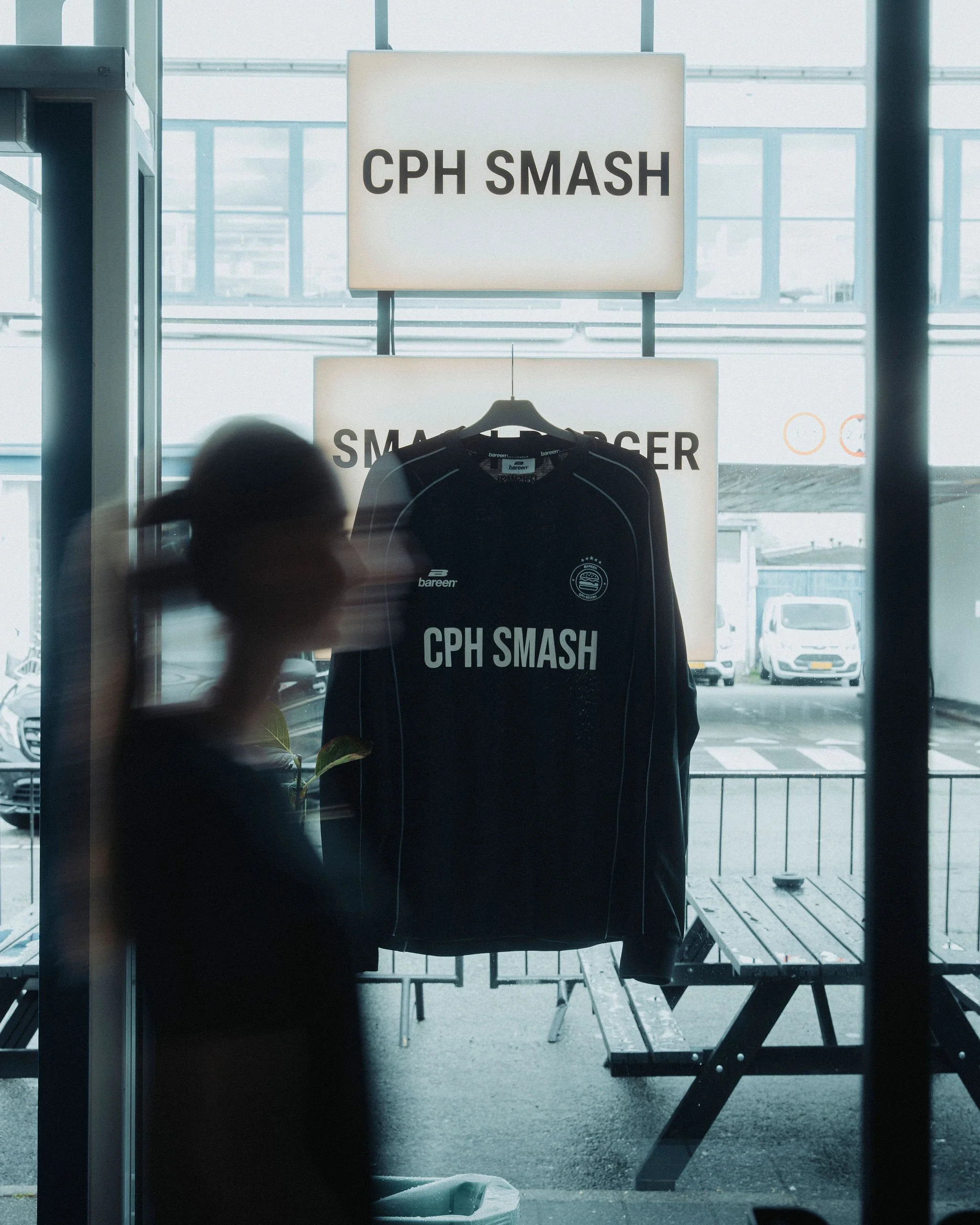 A retail display window with a 'CPH SMASH' jersey hanging on a hanger. The background shows an outdoor parking lot with several white cars and cloudy sky.