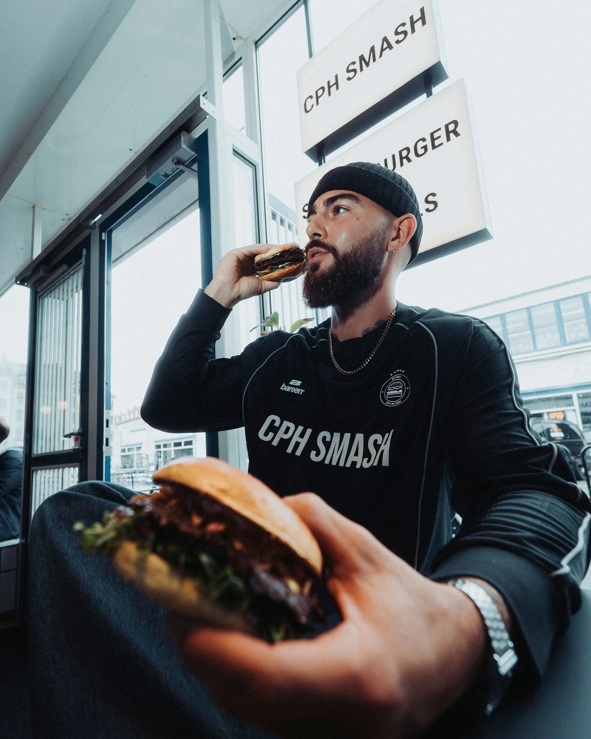 A man with a beard and earring, wearing a black beanie and black shirt, is sitting inside a restaurant or café, holding a burger in his right hand while looking away. The background shows a window and signage that reads 'CPH SMASH'.