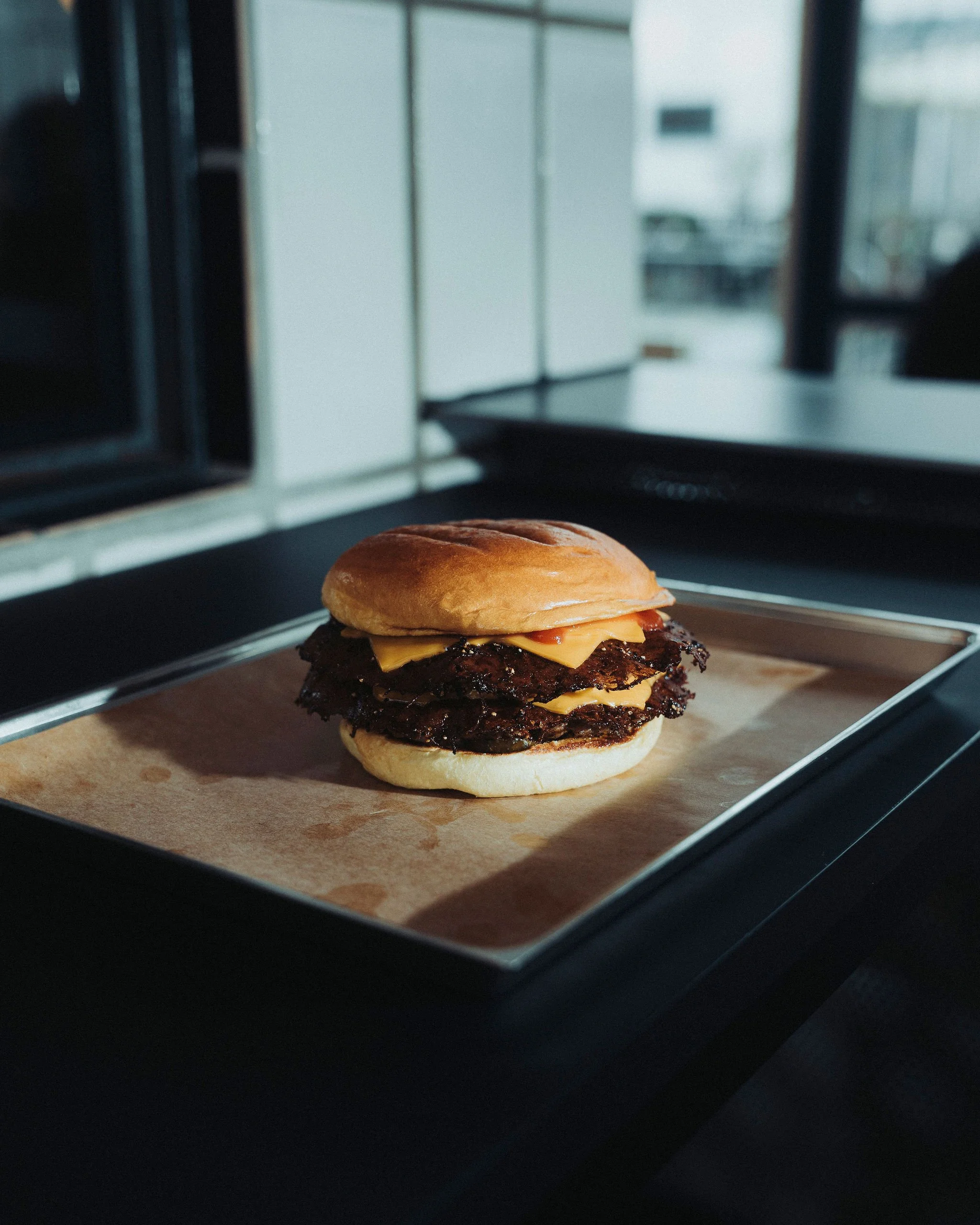 Close-up of a cheeseburger with a bun, melted cheese, a beef patty, and bacon, on a wooden surface with a blurry kitchen background.