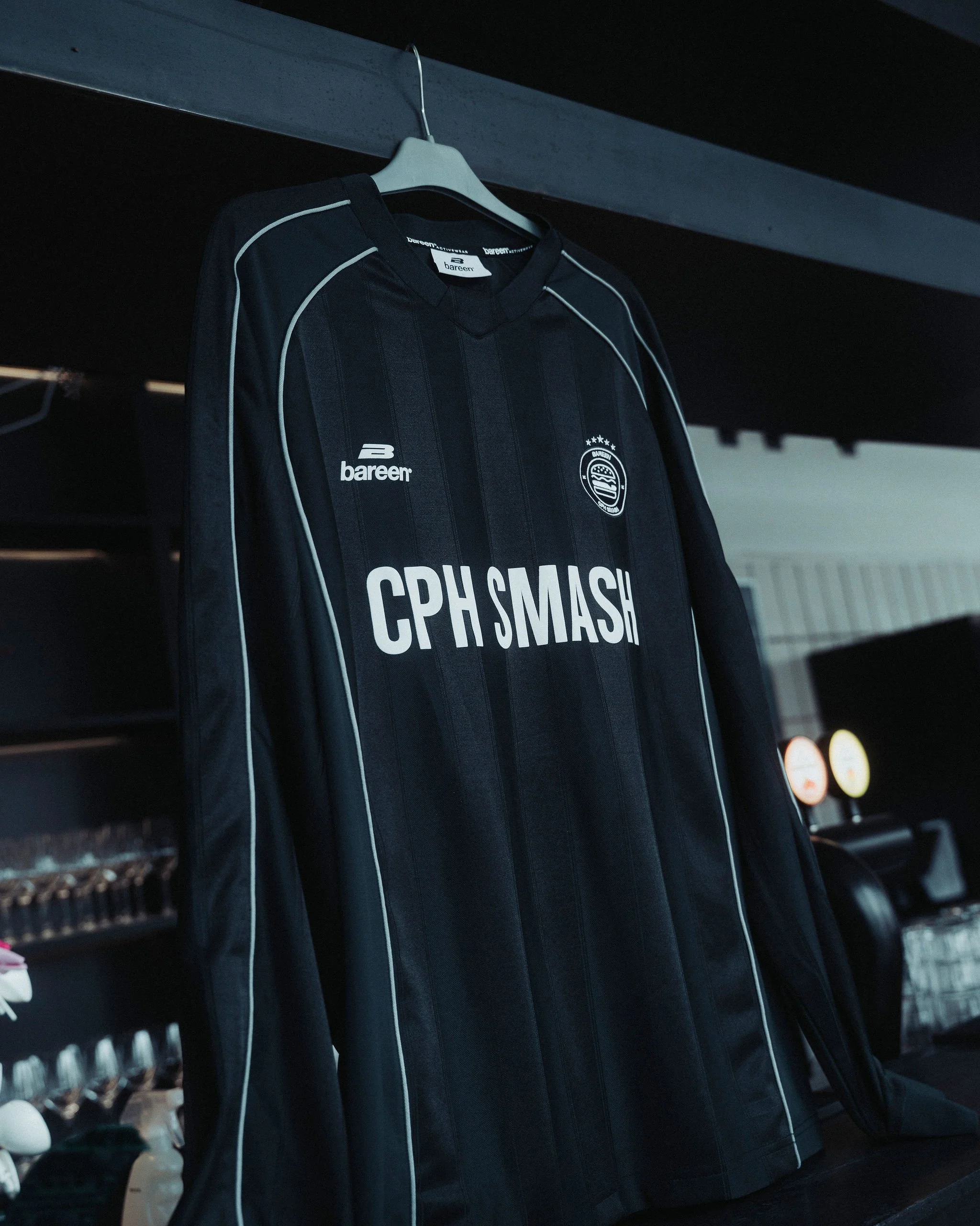 A black sports jersey with white piping hanging on a clothing hanger inside a closet, with the logo 'bareen' and the text 'CPH MASH' printed on the front.