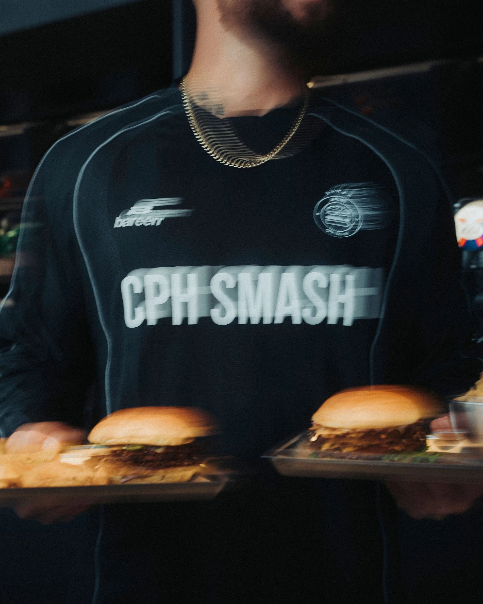 Blurred image showing a person wearing a black t-shirt with white text and graphics, and a gold chain necklace. The person is holding a tray with food items, including bread rolls, in the foreground.