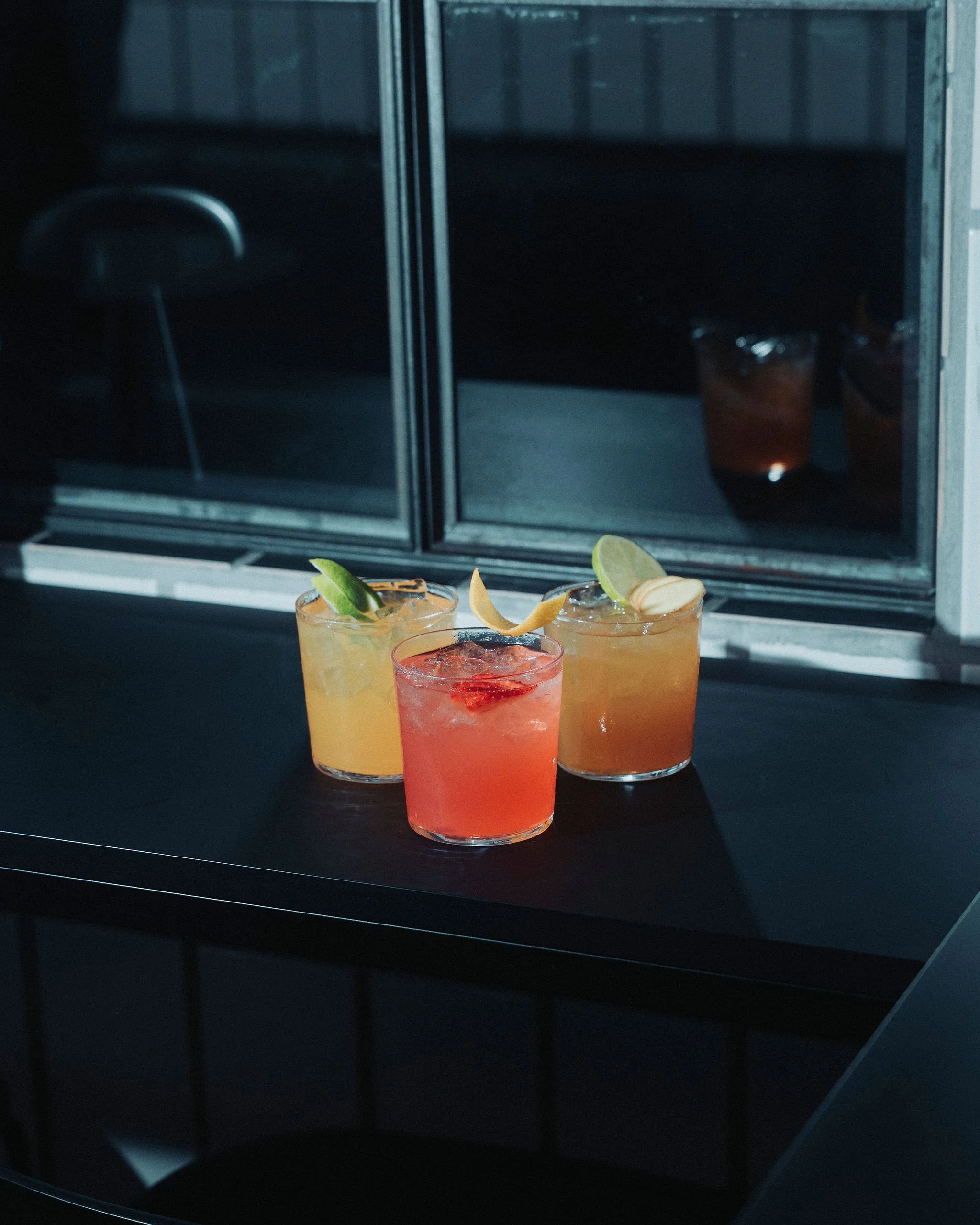 Three colorful cocktails with garnishes placed on a dark counter in front of a glass breakfast bar or window with a dark interior background.