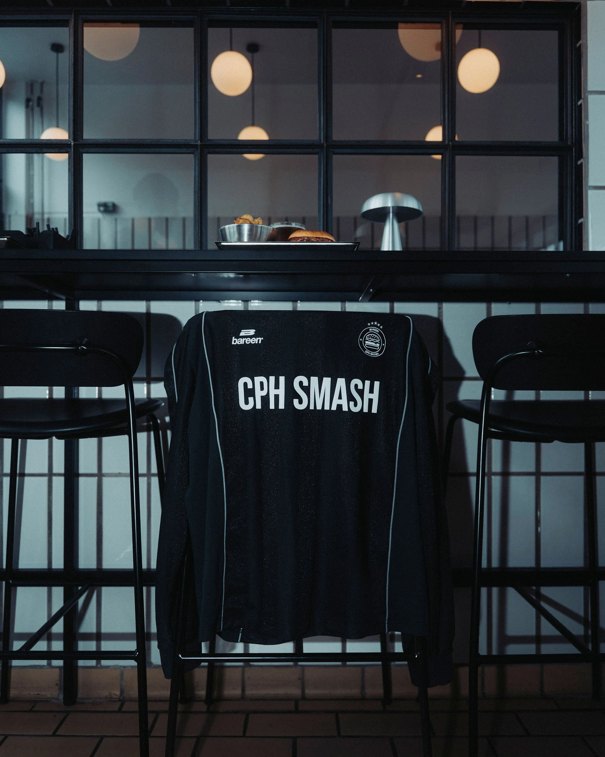 A black dining table with black chairs, with a black shirt hanging on one chair that has white CPH SMASH logo. Behind the table is a black partition with a glass window showing a counter with a metal bowl, a plate of food.