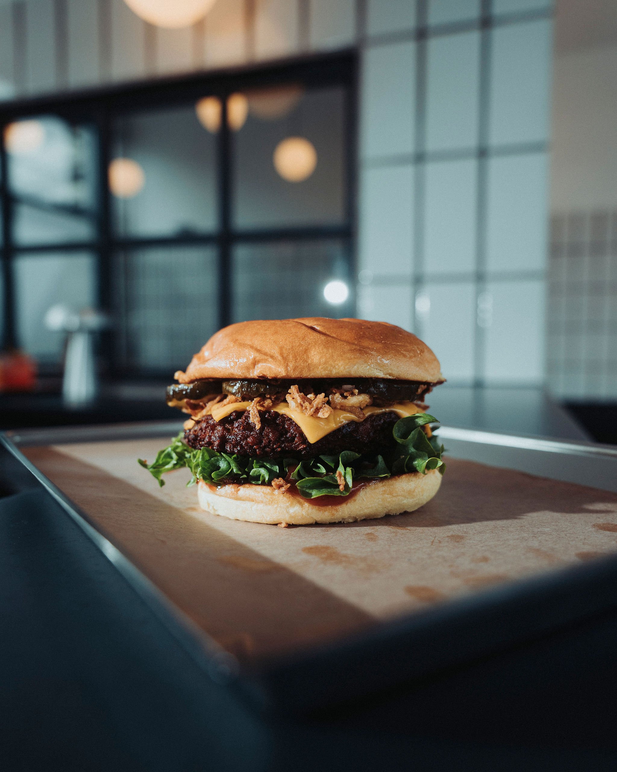 A close-up of a burger with a bun, lettuce, cheese, and a beef patty placed on a piece of parchment paper on a wooden surface, with the background showing part of a modern kitchen or restaurant interior.
