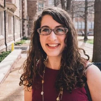 Young woman with curly brown hair wearing glasses and a maroon top outdoors in front of a brick building.