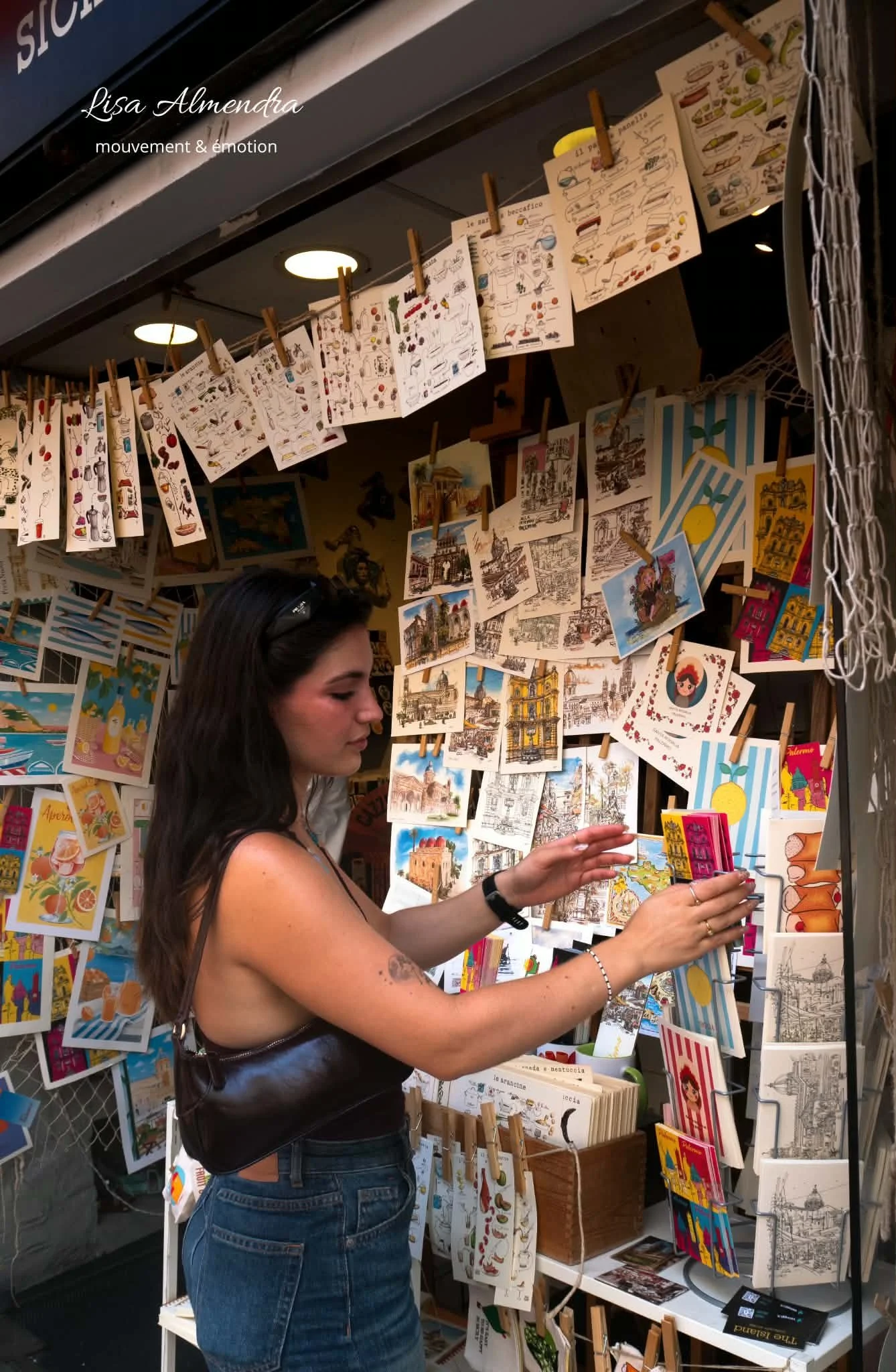 Femme achetant des cartes postales colorées dans un stand de marché. Le stand est décoré avec des cartes illustrées et des images encadrées, en plein air ou dans une boutique.