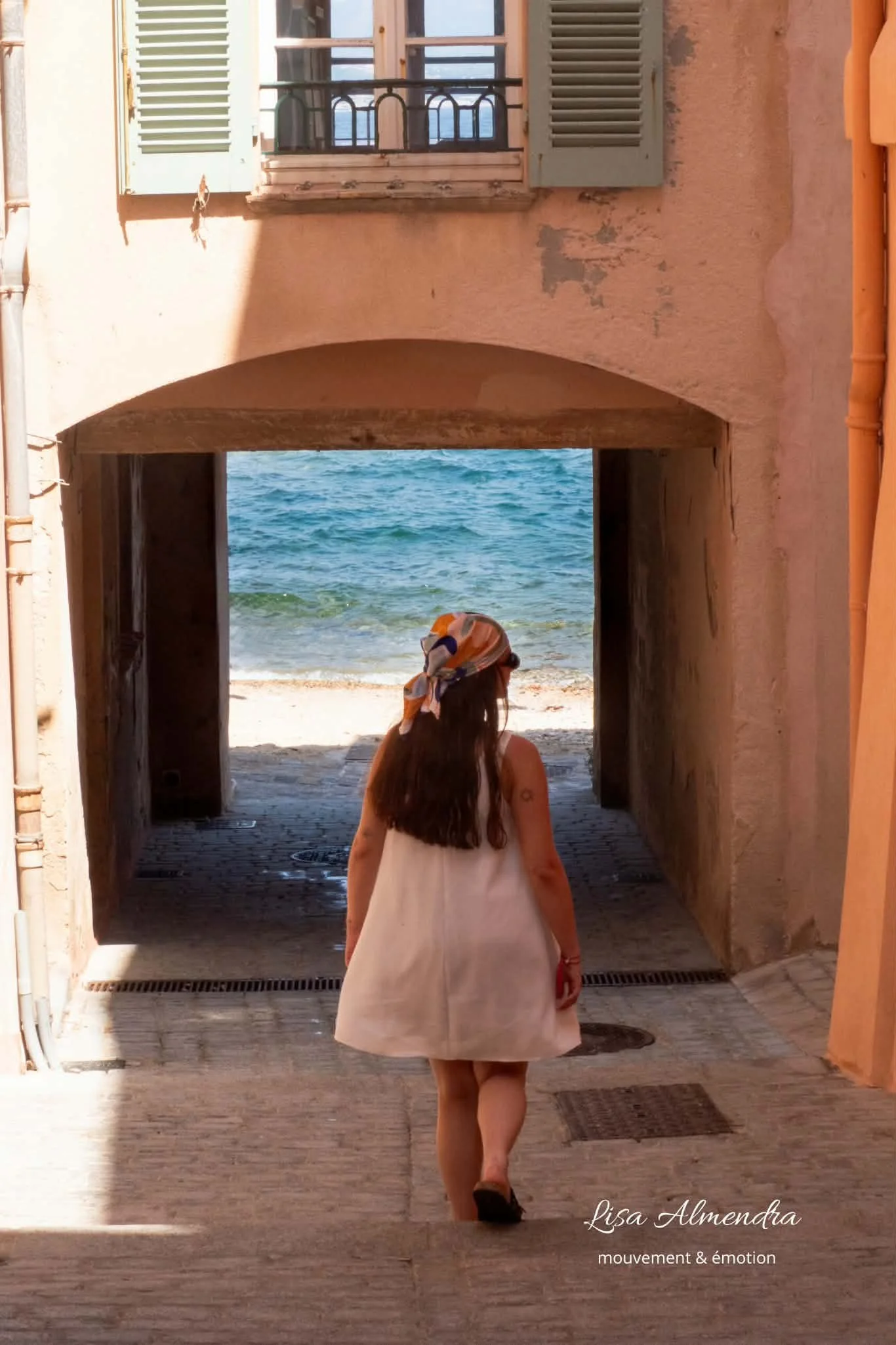 Une femme vêtue d'une robe blanc marche sous une arche vers la plage, avec la mer visible en arrière-plan. La lumière du soleil crée une ombre sur le mur à gauche. Sur le bas à droite, le texte indique : 'Lisa Almendra mouvement & émotion'.