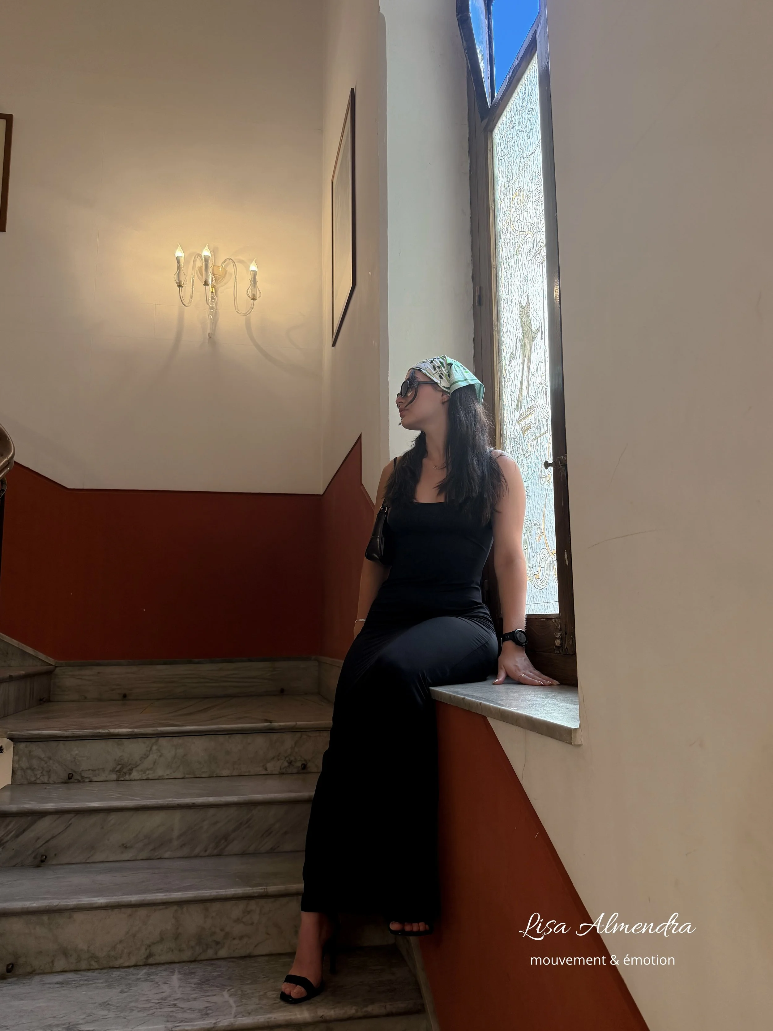 Une femme habillée en noir, portant des lunettes et un foulard, assise sur un rebord de fenêtre en marbre dans une pièce avec des murs beige, un mur rouge et un lustre à quatre ampoules