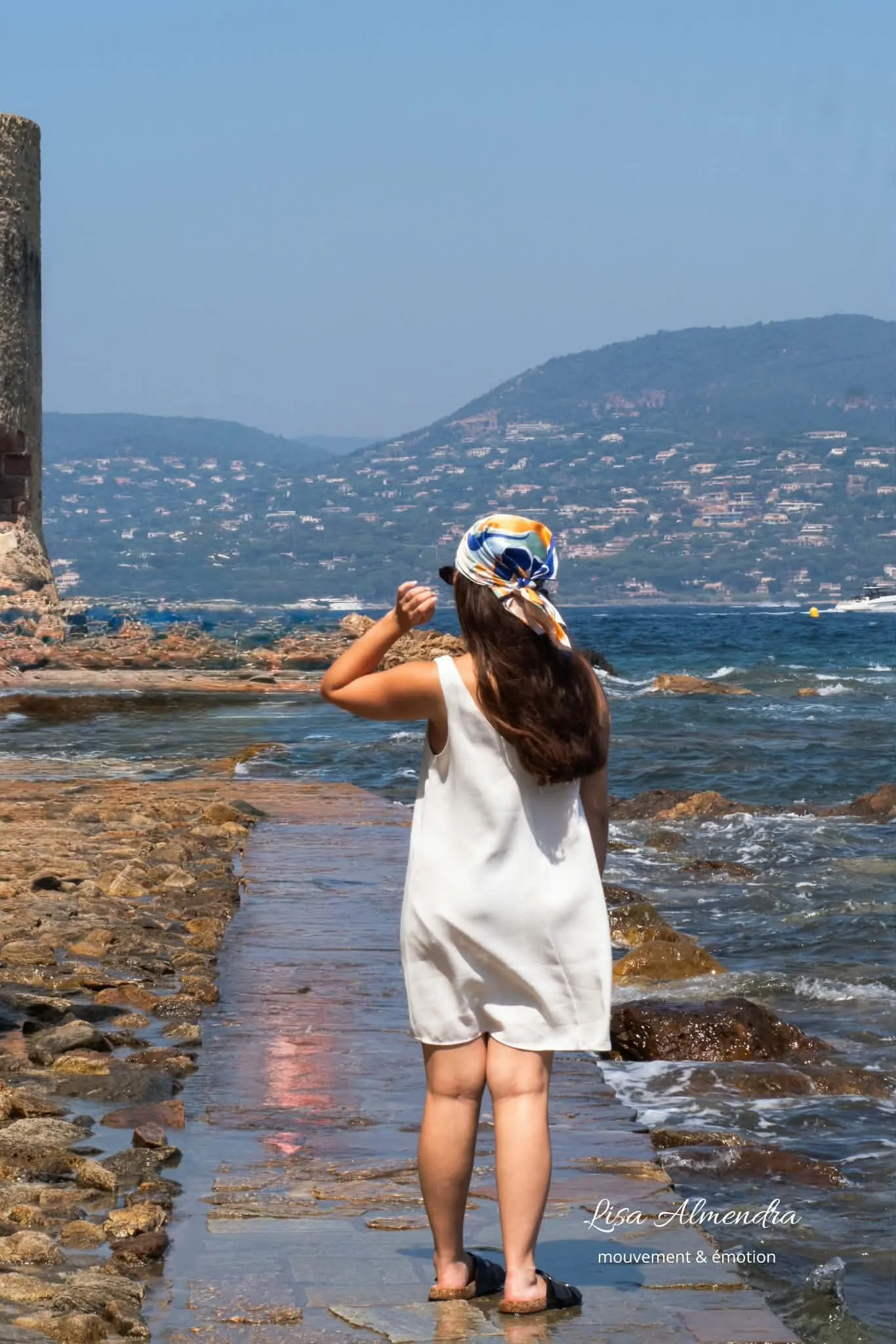 Jeune femme portant une robe blanche, un foulard coloré dans les cheveux, debout sur une plage rocheuse avec vue sur la mer et des collines au fond, en été.