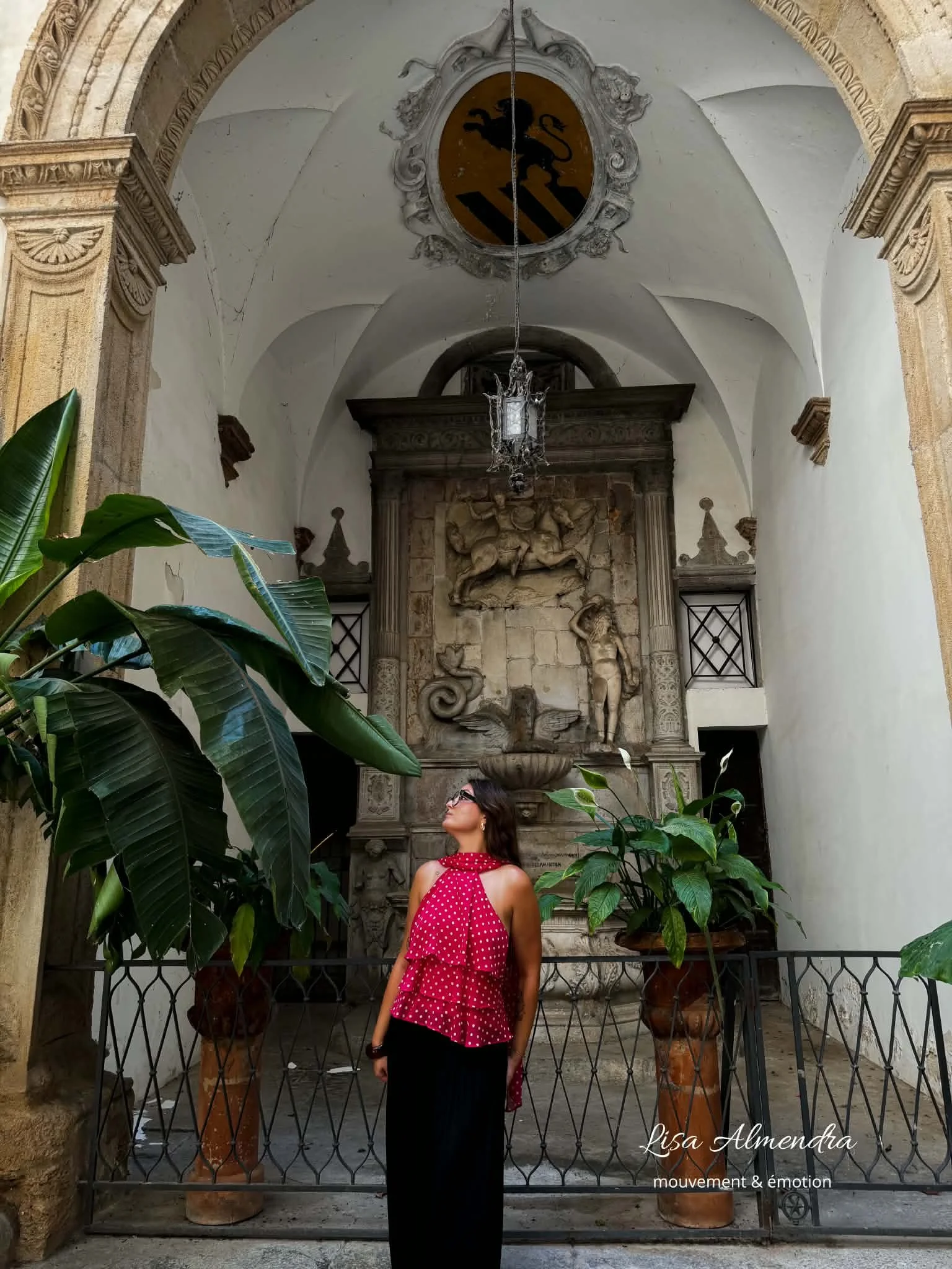 Une femme portant une robe rouge à pois blancs se tenant devant une grille en fer forgé avec une fontaine et une sculpture antique en pierre derrière elle dans un bâtiment historique, décoré avec des plantes vertes et un plafond voûté.