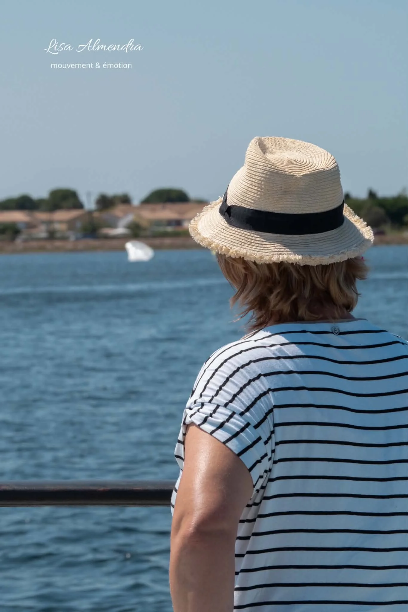 Une personne portant un chapeau en paille à rayures noires et blanches regarde une étendue d'eau, avec des bâtiments en arrière-plan et un ciel clair.