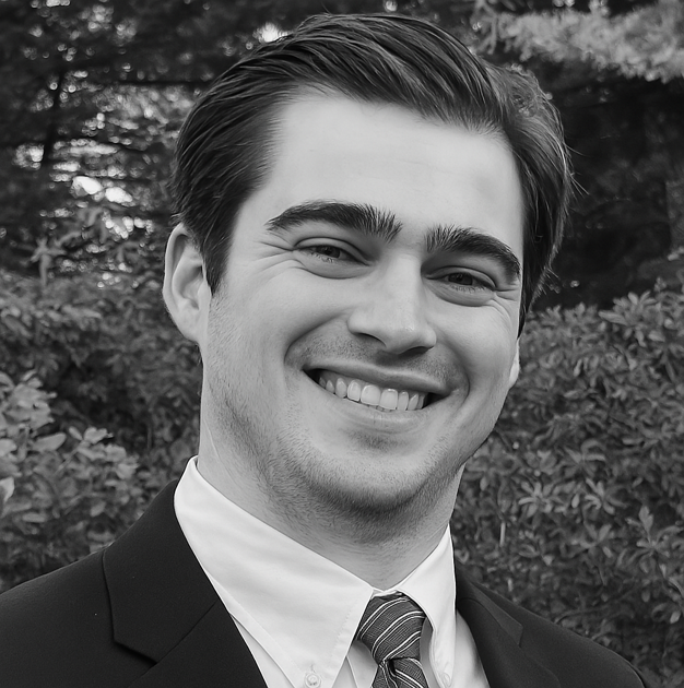 Portrait of a young man in a suit, smiling, with trees and foliage in the background.