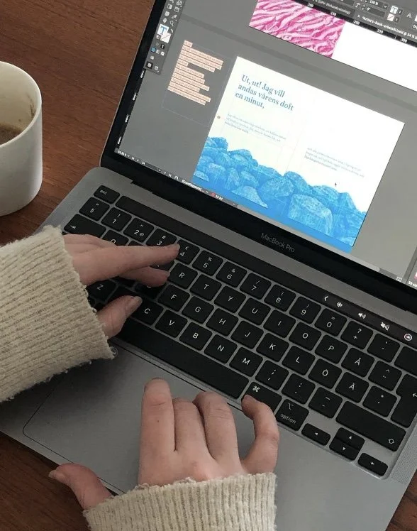 Person som arbetar på en MacBook Pro, med en designprogramvara öppen på skärmen, samt en kopp kaffe på ett träbord.
