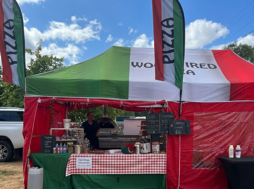 A pizza stand at an outdoor event with a green, white, and red canopy, flags, and a chalkboard menu. The stand sells cheese and other flavored pizzas for six pounds. A woman is working behind the counter, with a pizza oven and toppings visible.