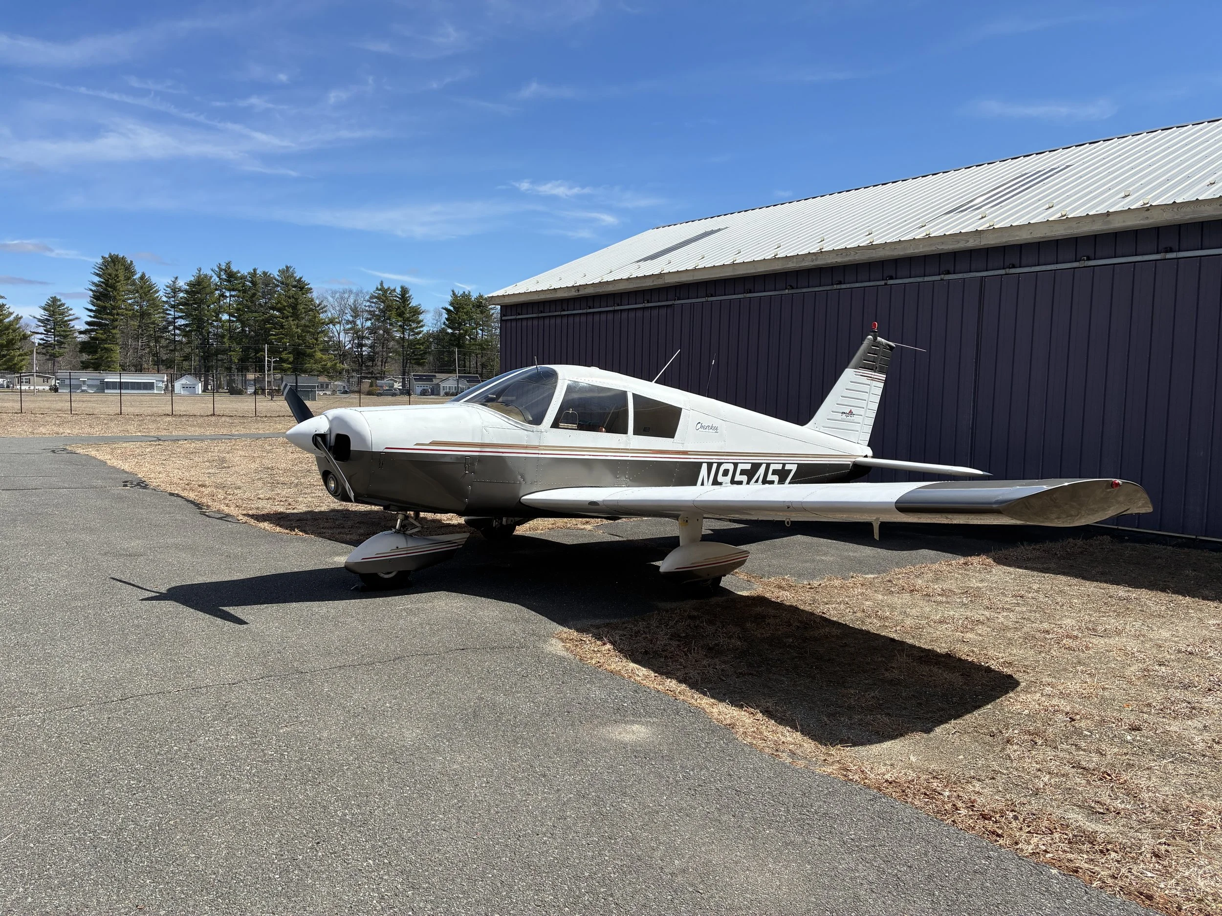 A white small aircraft with the registration N94547 parked on a tarmac area next to a dark-colored building with a metal roof. There are trees and houses in the background under a blue sky with some clouds.