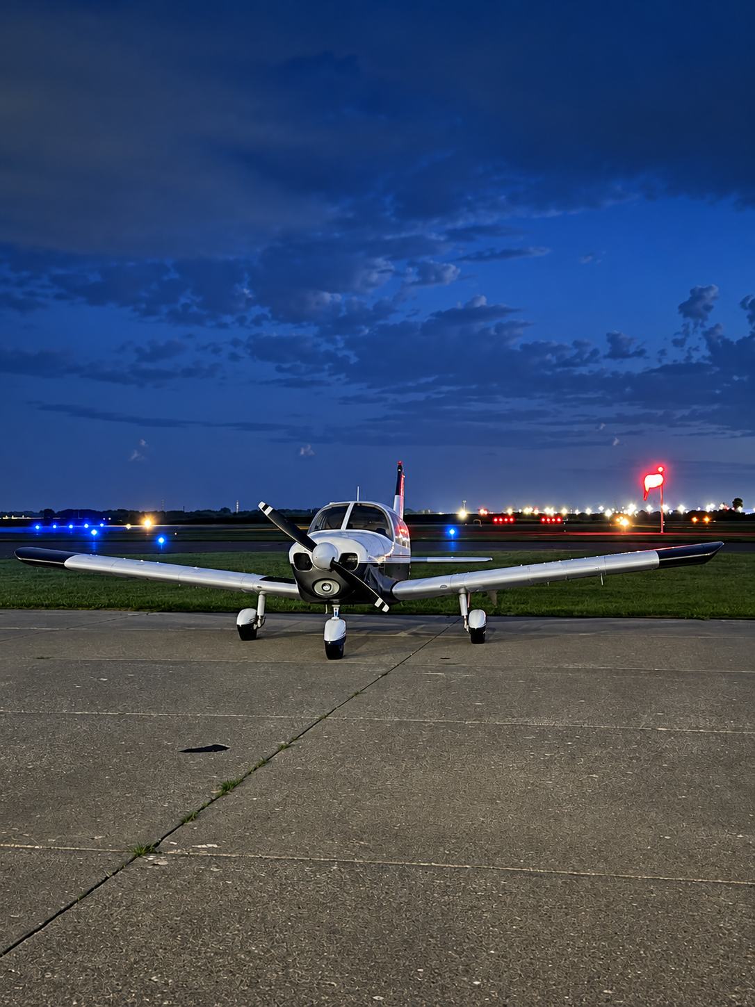 Small private airplane parked on an airport tarmac at dusk with a dark blue sky, runway lights, and illuminated windsocks in the background.